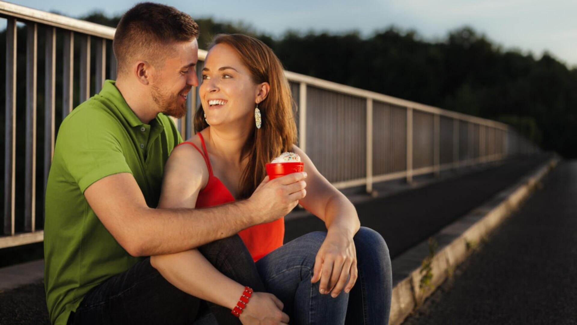 A smiling couple sits close together on a sidewalk near a railing, with the woman holding a red cup of ice cream. The man wears a green shirt and the woman wears a red top, both looking happy and affectionate.