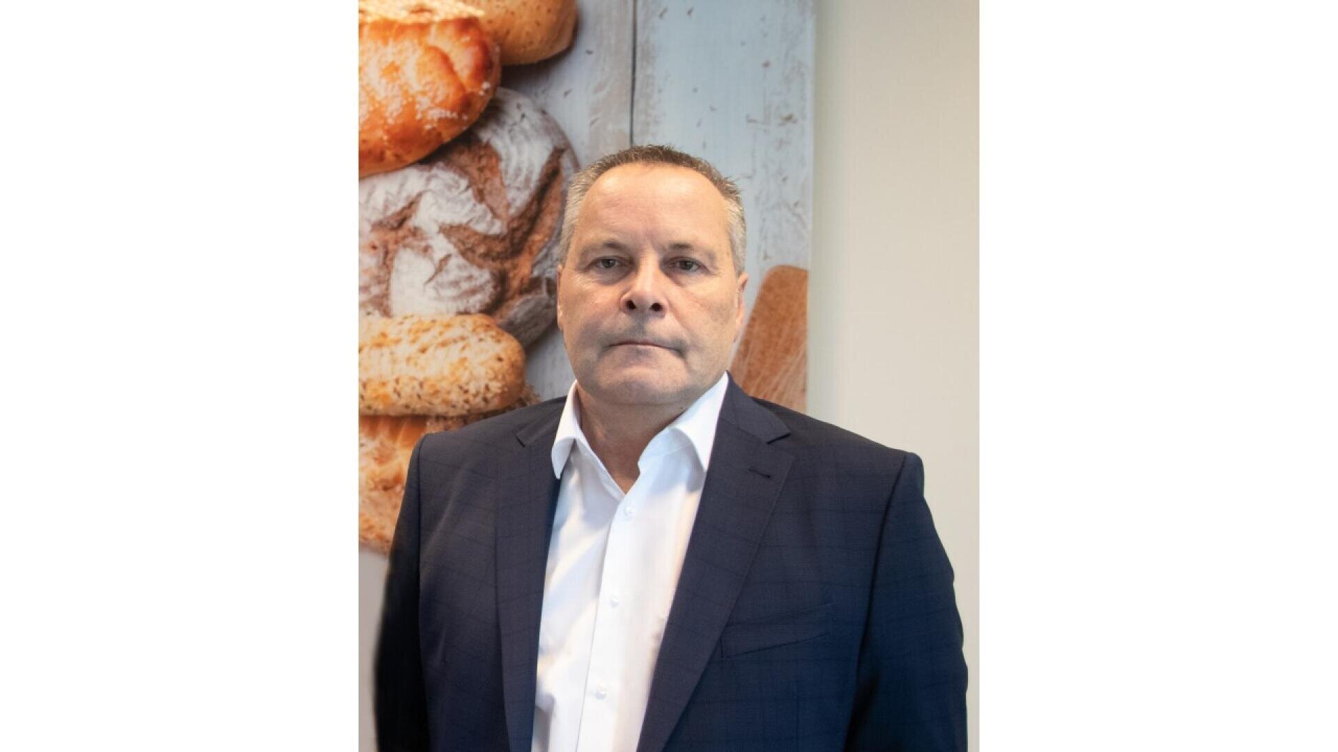 A man in a dark suit and white shirt stands indoors in front of a wall with images of assorted bread loaves. He looks directly at the camera with a neutral expression.