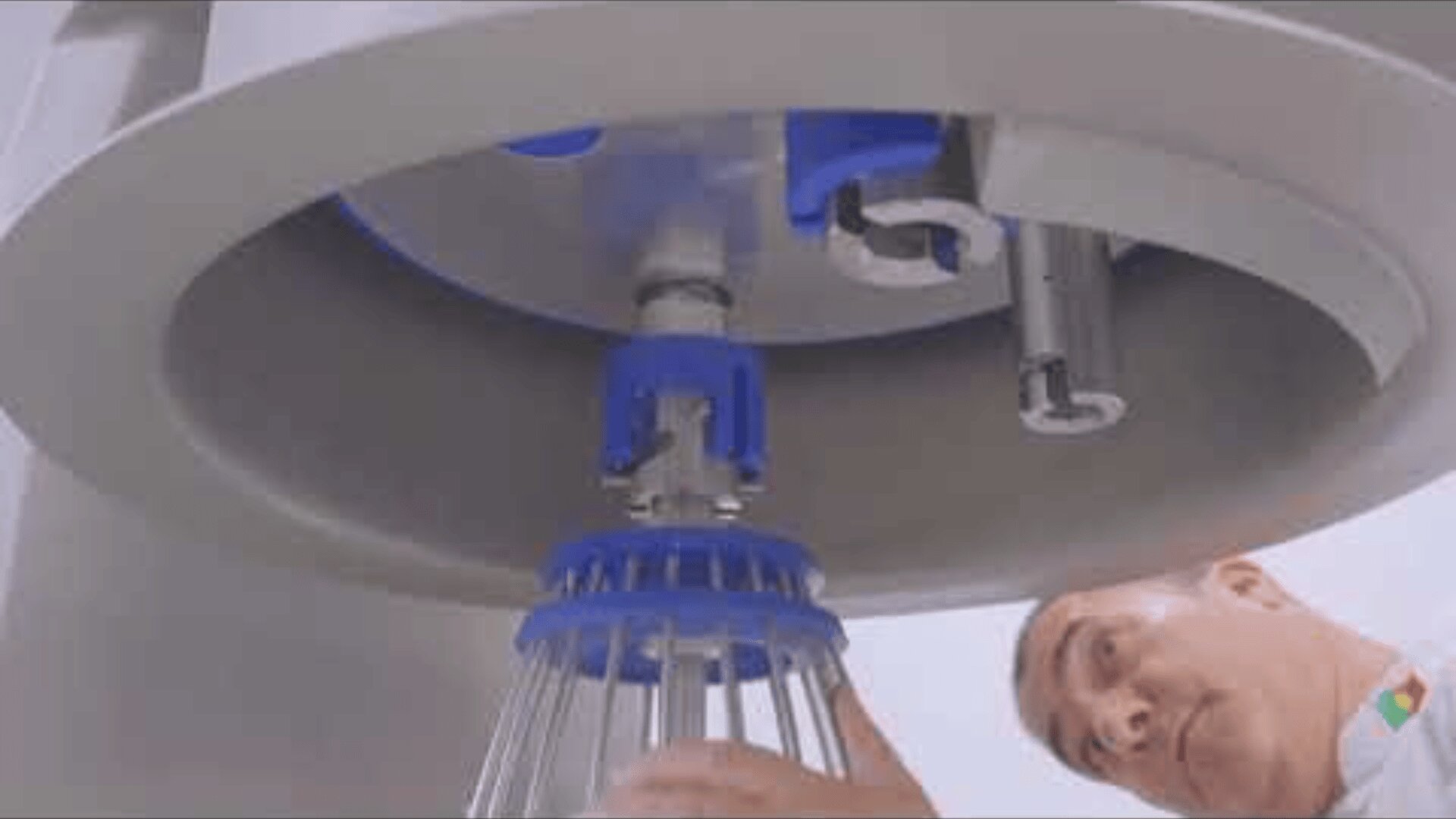 A person examines the underside of a large industrial machine and focuses on blue and metal mechanical parts reminiscent of a whisk.