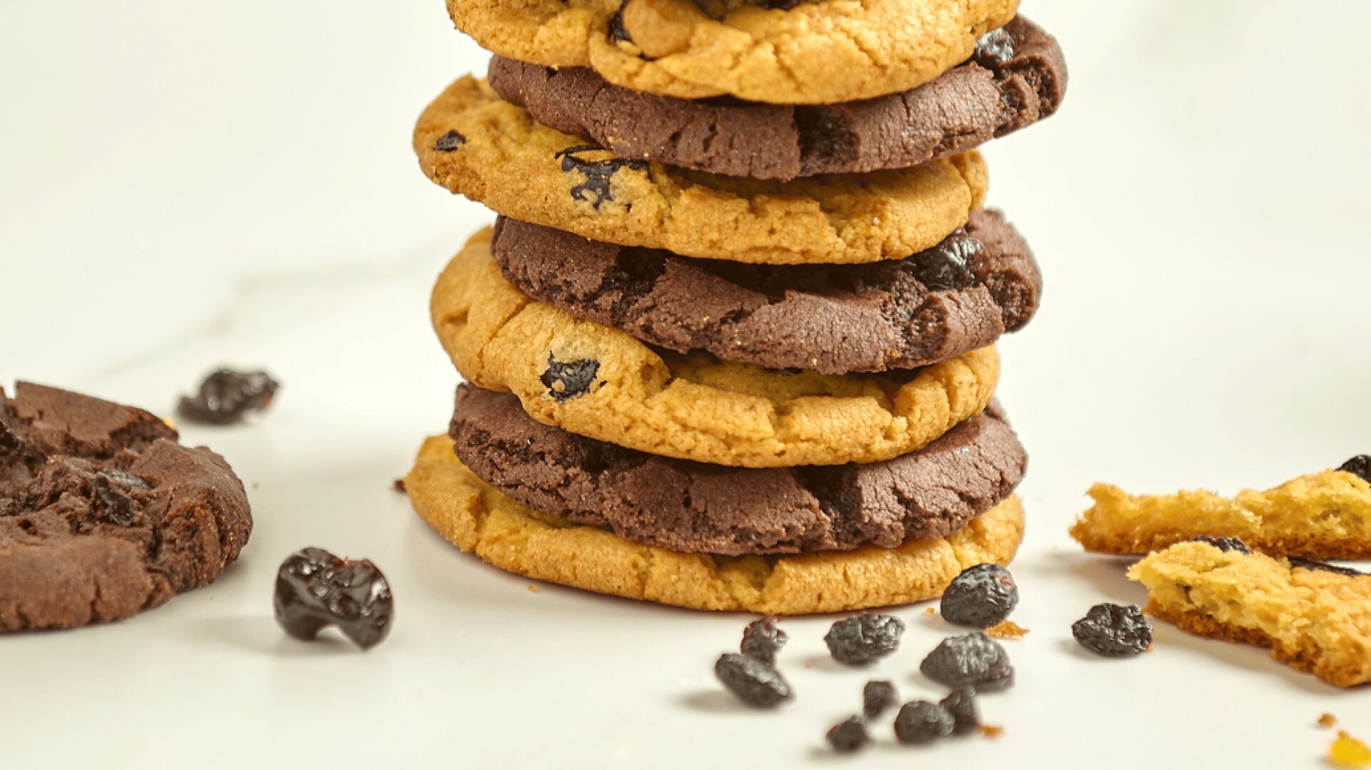 A stack of alternating chocolate and plain cookies with dried fruit sits on a white surface. Beside the stack are a broken cookie, scattered dried fruit, and cookie crumbs.