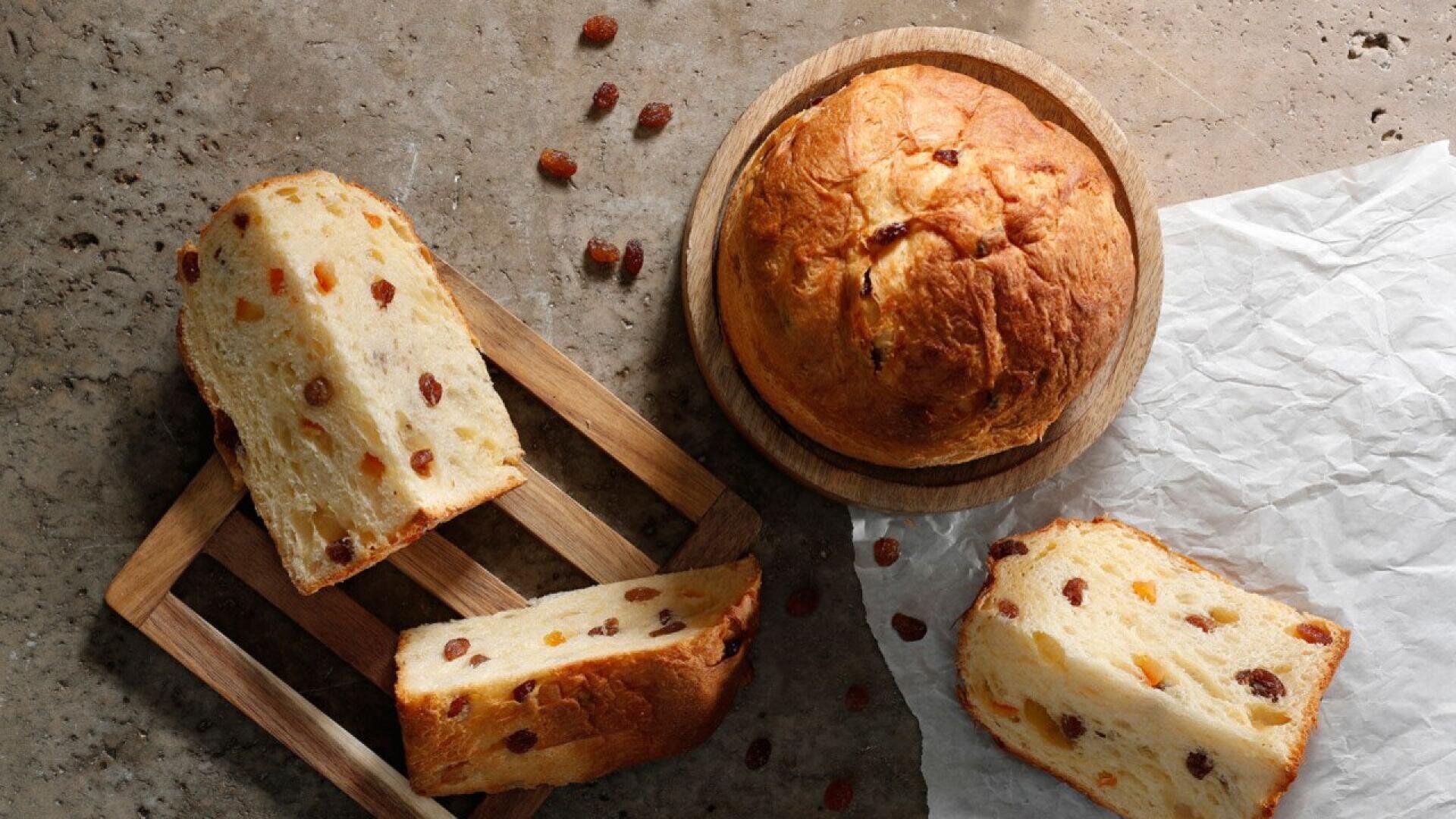 Overhead view of a round loaf of panettone bread on a wooden plate, with slices of panettone showing bits of raisins, placed on a wooden tray and parchment paper, on a textured stone surface.