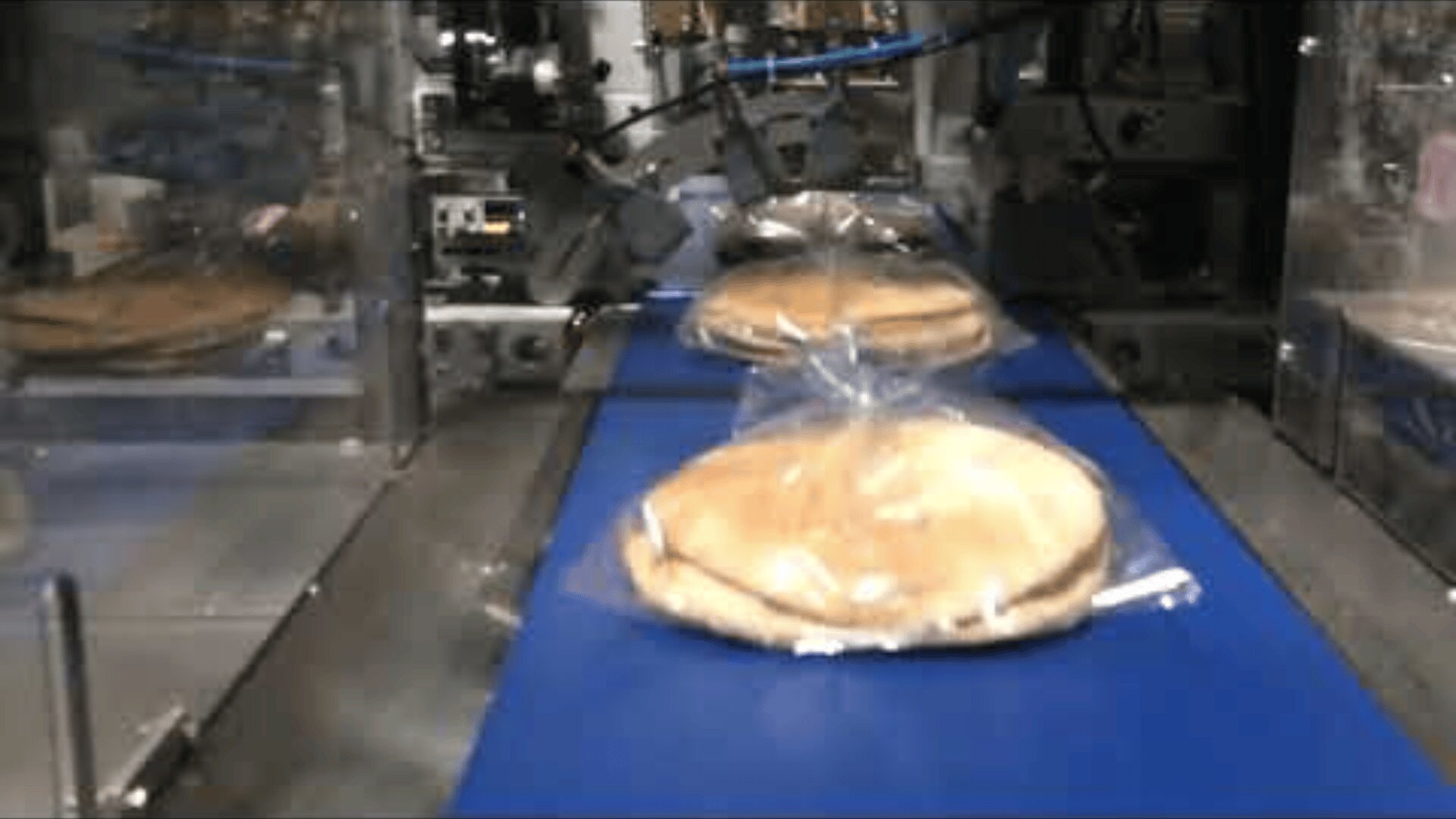 Stacks of pita bread, wrapped in clear plastic, move along a blue conveyor belt inside a food processing and packaging facility.