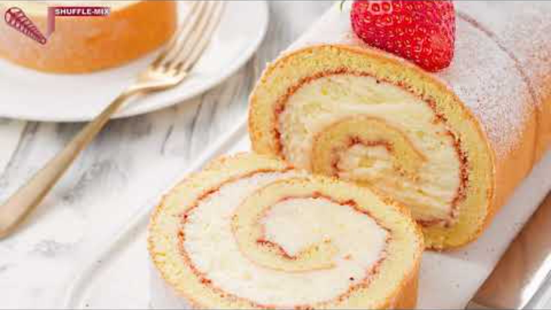 Close-up of a sliced Swiss roll cake with a creamy swirl filling, topped with a fresh strawberry, on a white marble surface next to a fork and plate.