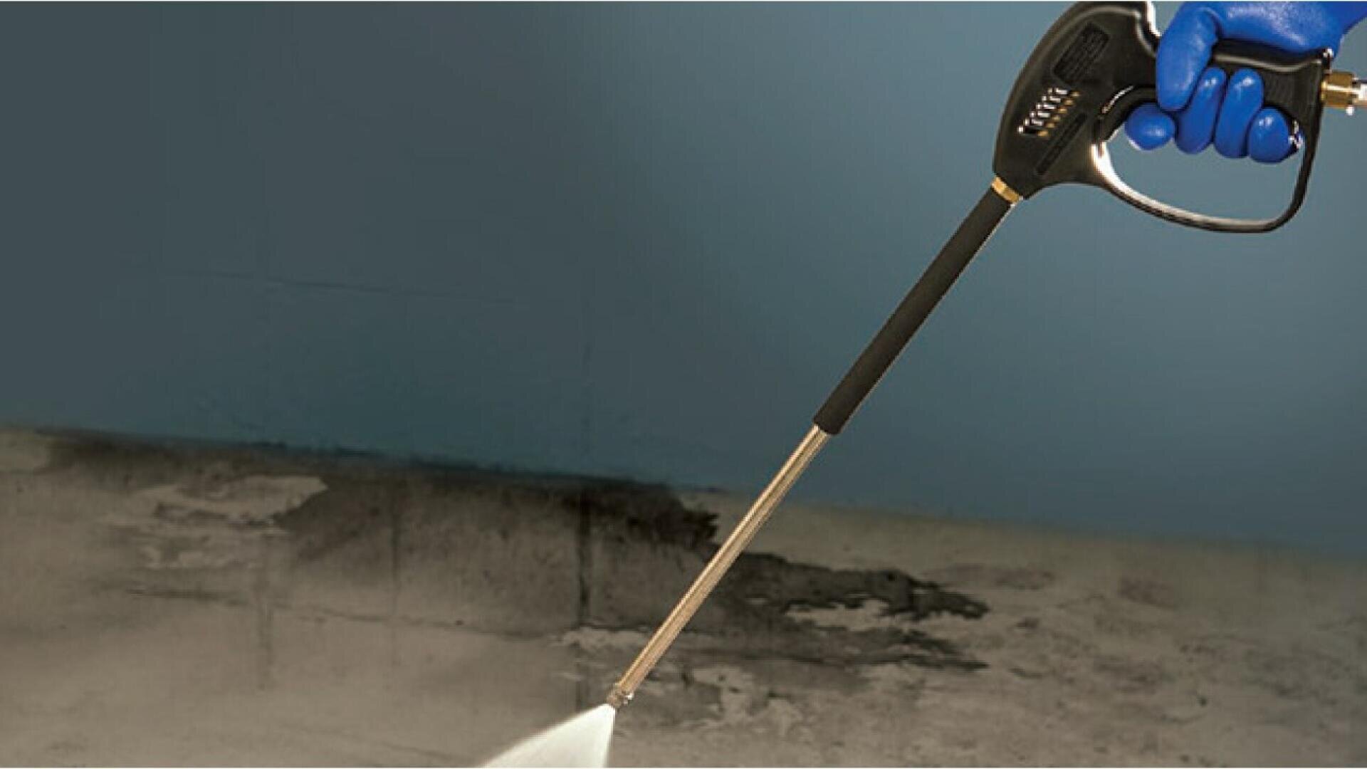 A person wearing a blue glove uses a pressure washer to clean a dirty concrete surface, showing a clear contrast between the cleaned and uncleaned areas.