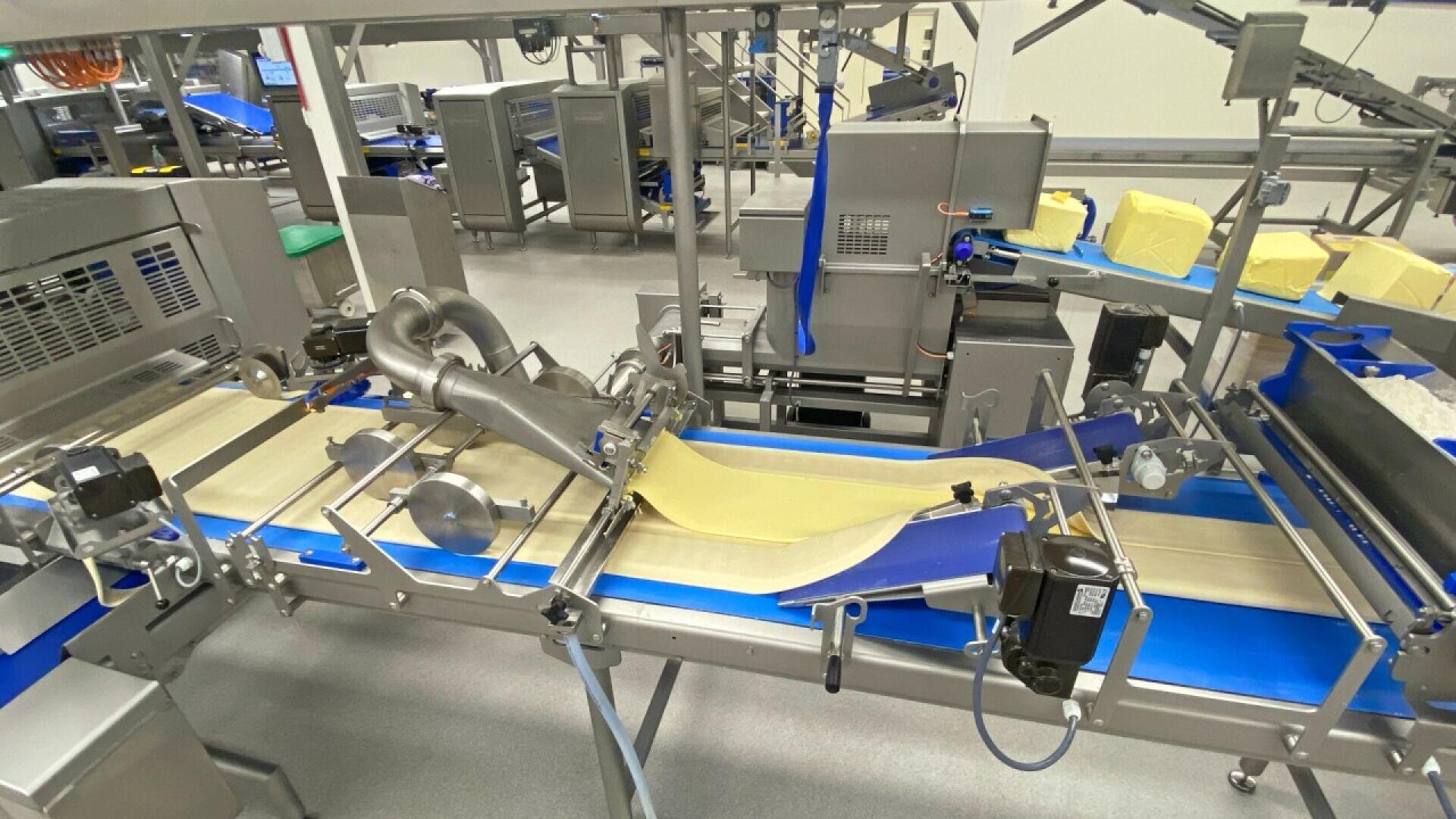 Eine Lebensmittelverarbeitungsmaschine in einer Fabrik formt und transportiert Teigplatten oder Gebäck auf blauen und beigen Förderbändern, während im Hintergrund gelbe Butter- oder Margarineblöcke zu sehen sind.