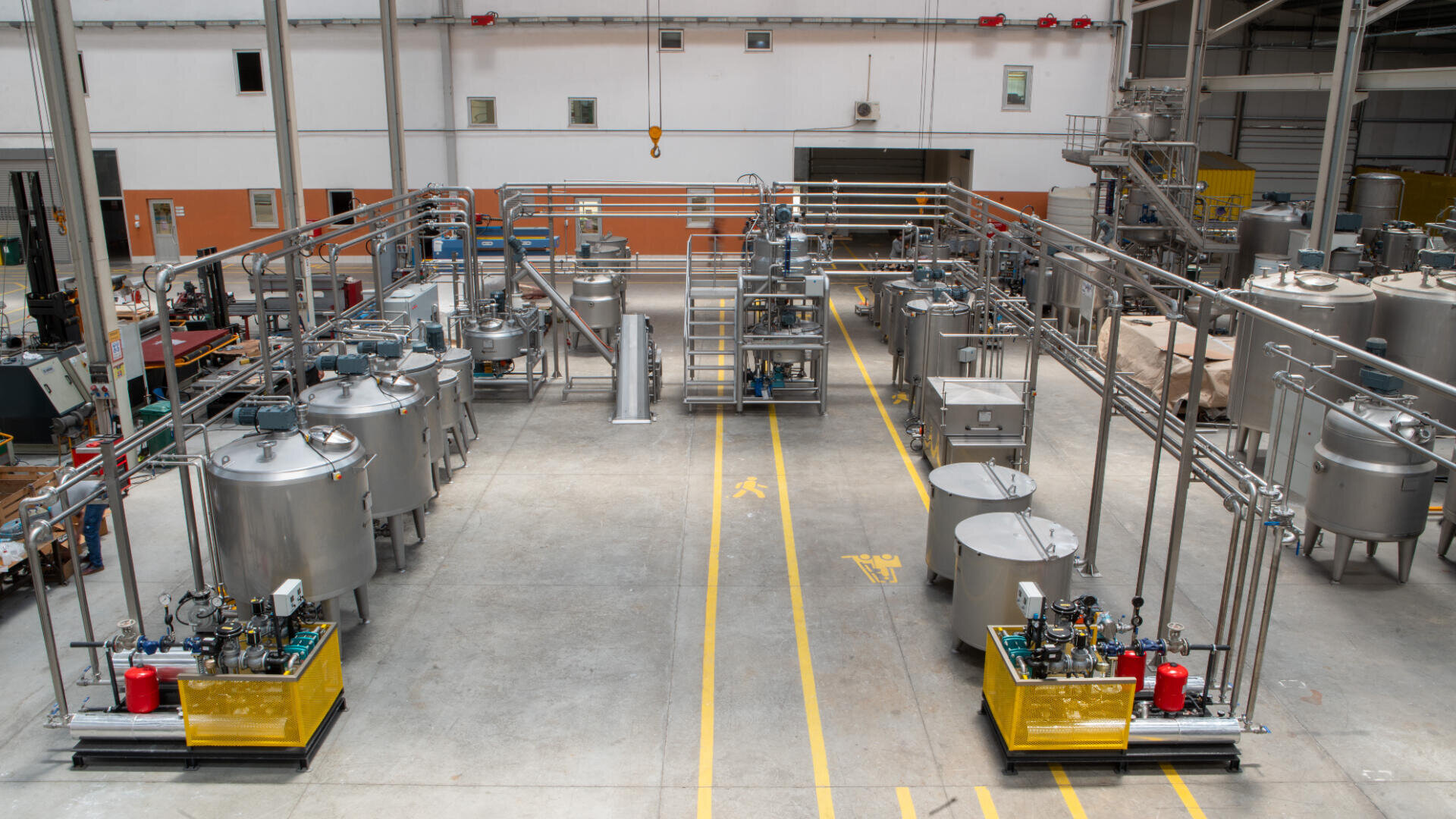 Large industrial facility with multiple stainless steel tanks, pipes, and machinery arranged in rows on a clean, spacious factory floor with yellow safety lines and high ceilings.