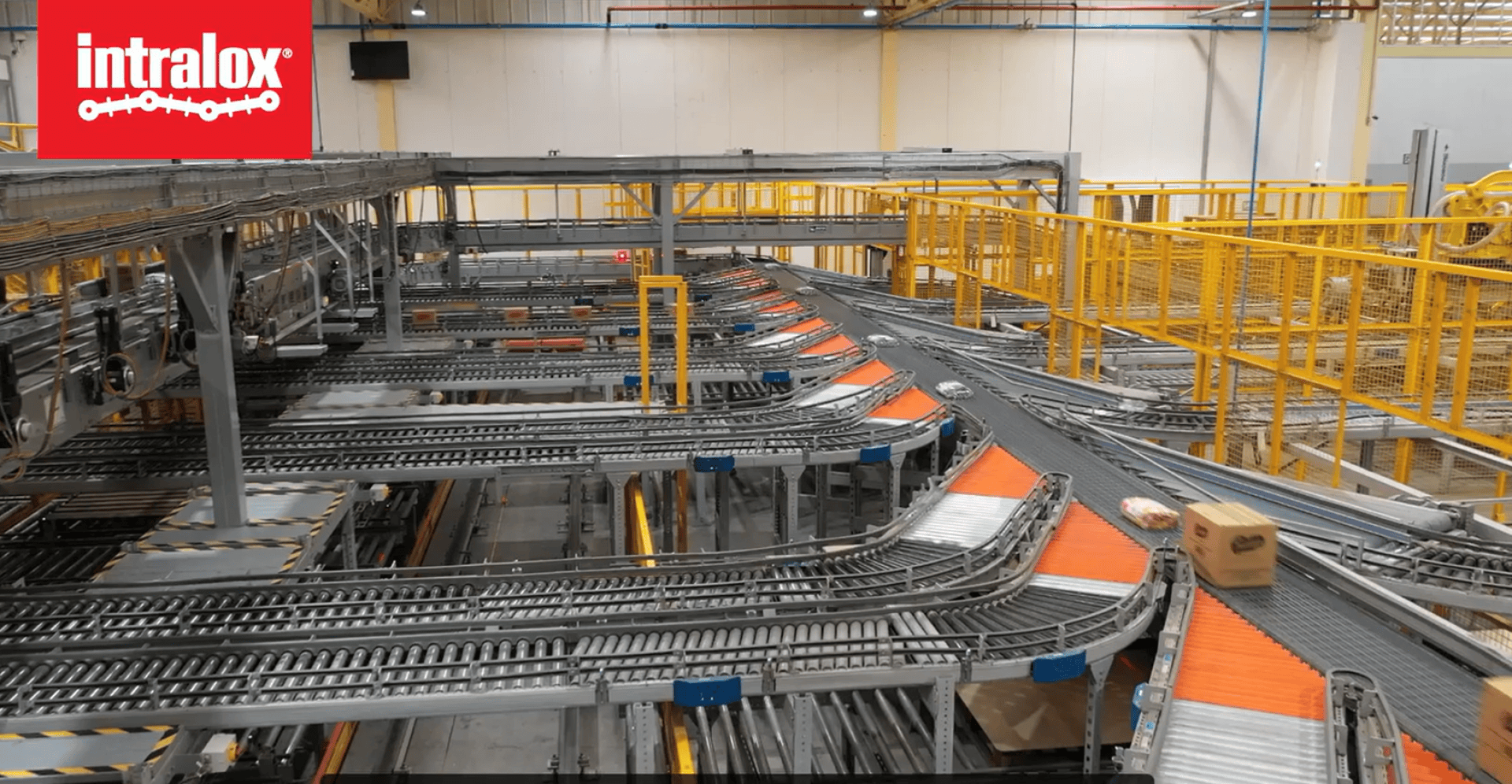 A modern conveyor system with orange and gray sections sorts packages in a large industrial facility. The Intralox logo appears in the top left corner. Yellow safety railings line the area.