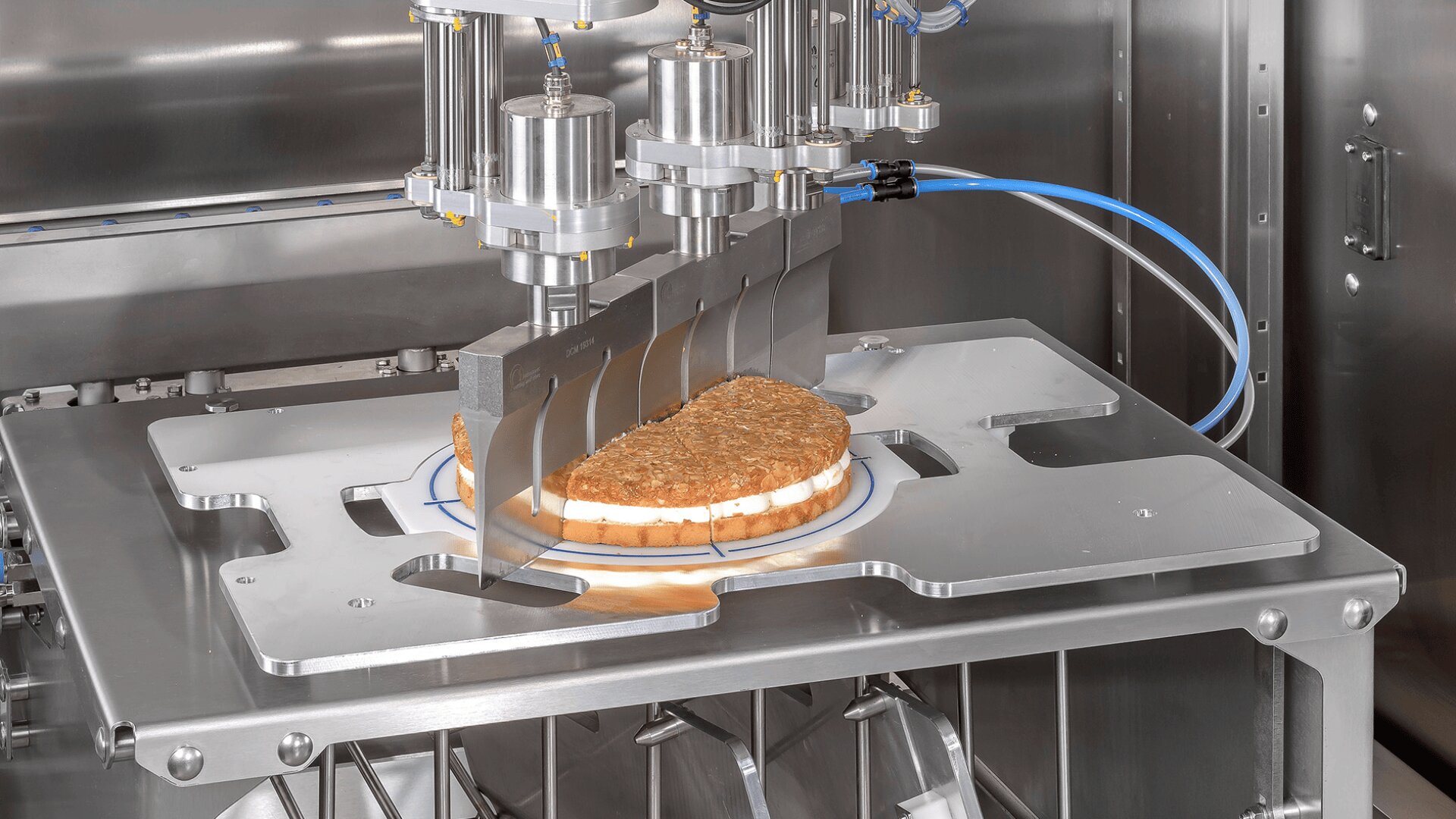 Nahaufnahme einer industriellen Lebensmittelverarbeitungsmaschine, die ein rundes, geschichtetes Sandwich oder einen Kuchen auf einem Metalltablett schneidet, wobei die mechanischen Arme und Klingen in einer sauberen Edelstahlumgebung sichtbar sind.