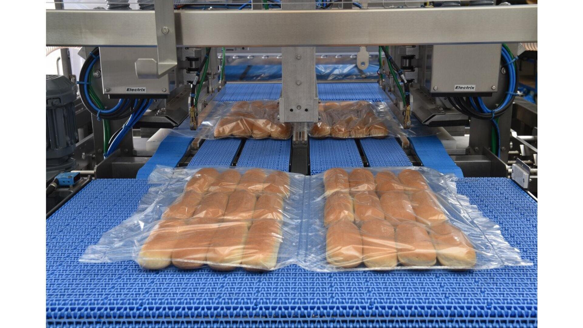 Four packages of hot dog buns in clear plastic are on a blue conveyor belt, moving through a food processing machine in an industrial bakery setting.