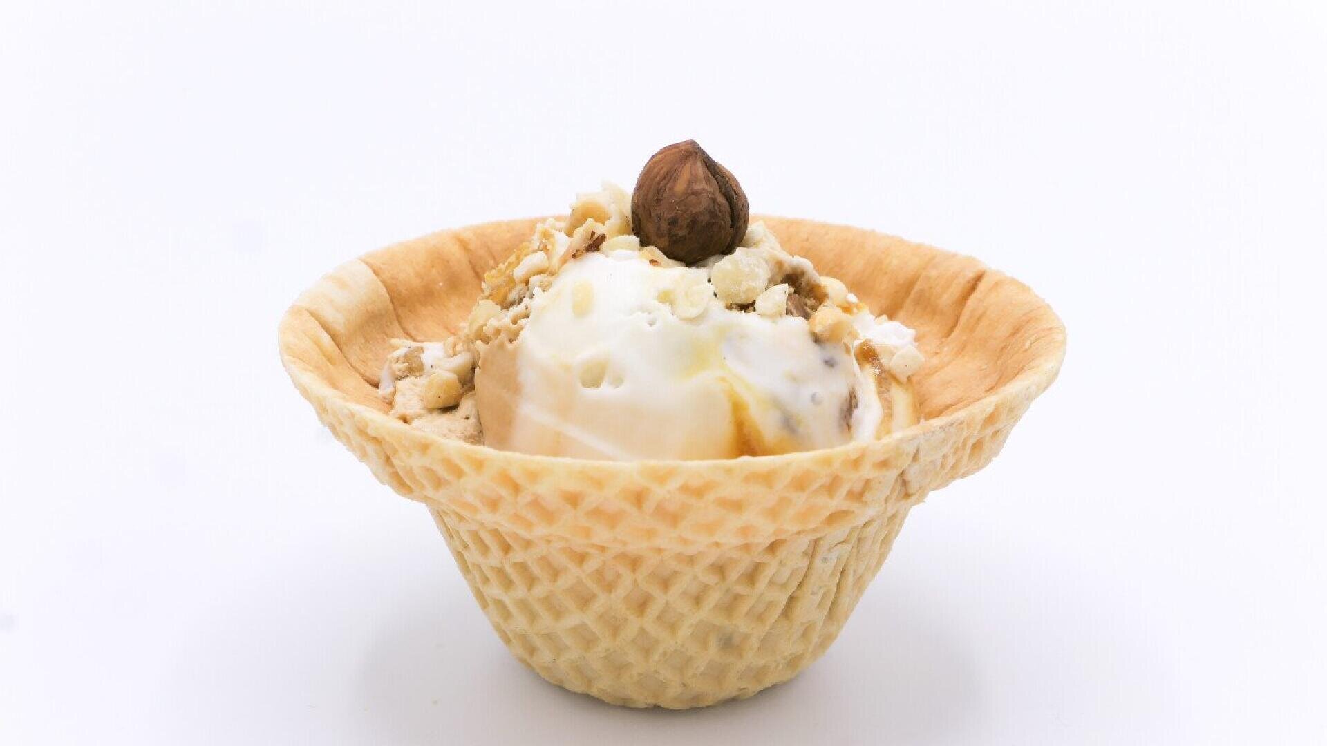 A scoop of vanilla ice cream in a waffle bowl, topped with a whole hazelnut and crushed nuts, on a white background.