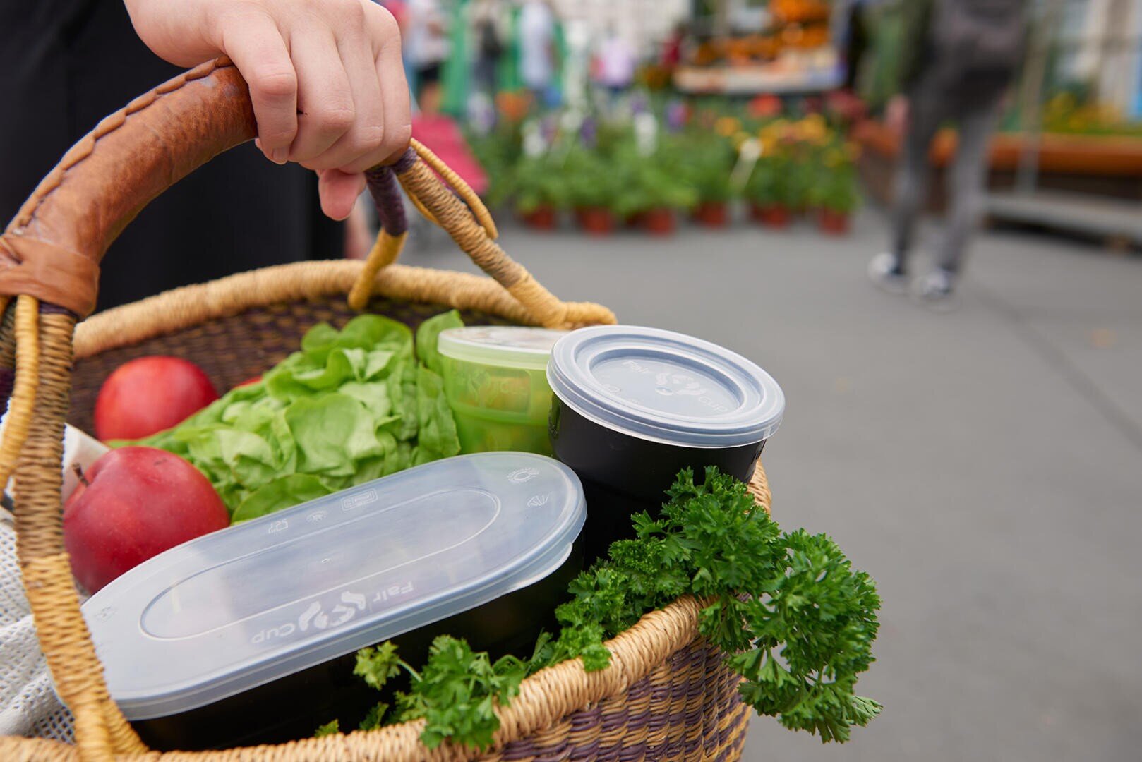 Eine Hand hält einen geflochtenen Korb, der mit frischem Gemüse, Äpfeln, Petersilie und wiederverwendbaren Lebensmittelbehältern gefüllt ist, auf einem Markt im Freien mit unscharfen Pflanzen und Menschen im Hintergrund.
