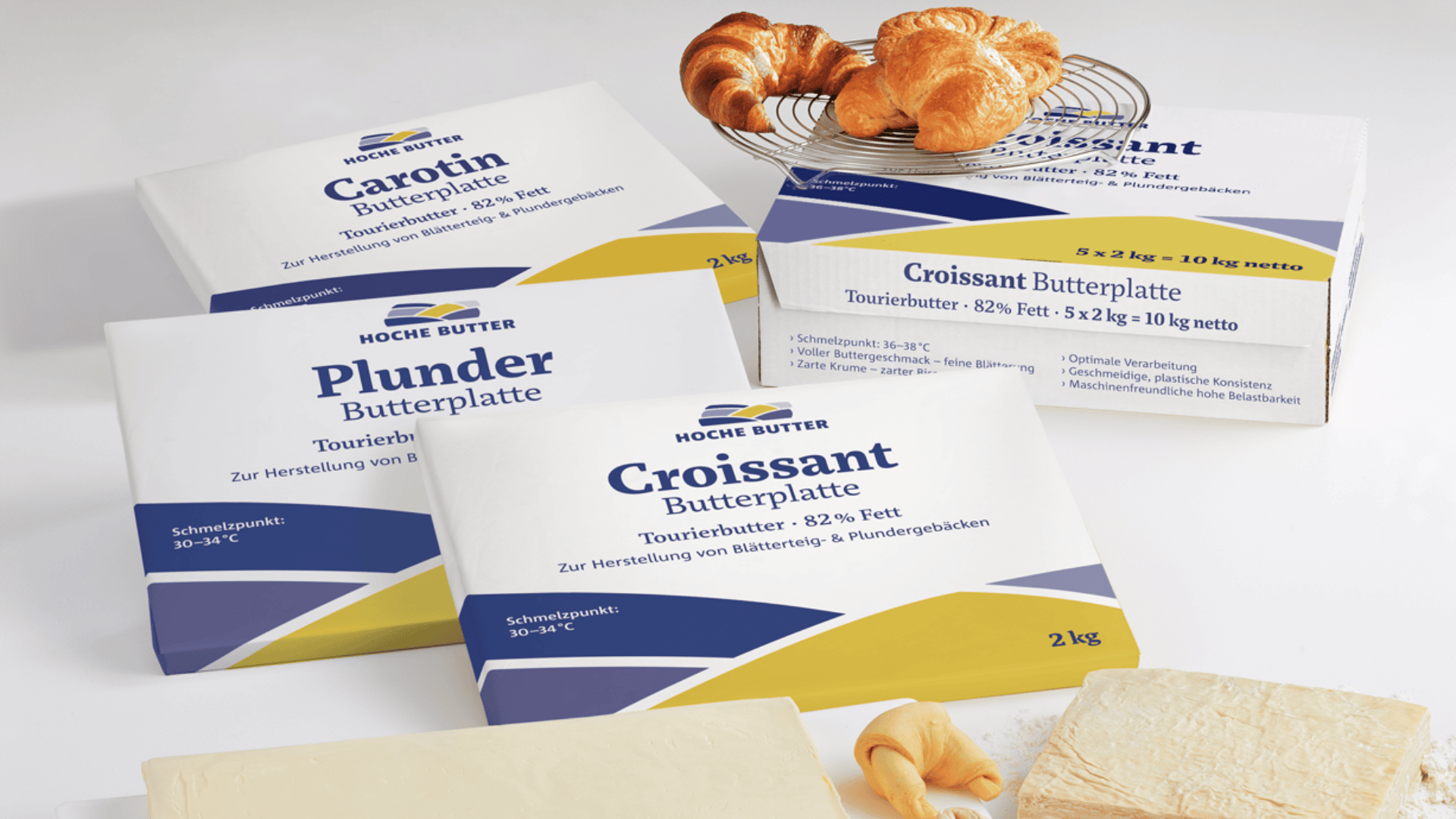 Vier Verpackungen von Hoche Butterprodukten mit den Bezeichnungen Croissant, Carotin und Plunder Butterplatte werden mit Butterblöcken und drei Croissants auf einer weißen Fläche präsentiert. Die Verpackung ist überwiegend weiß mit blauen und gelben Akzenten.