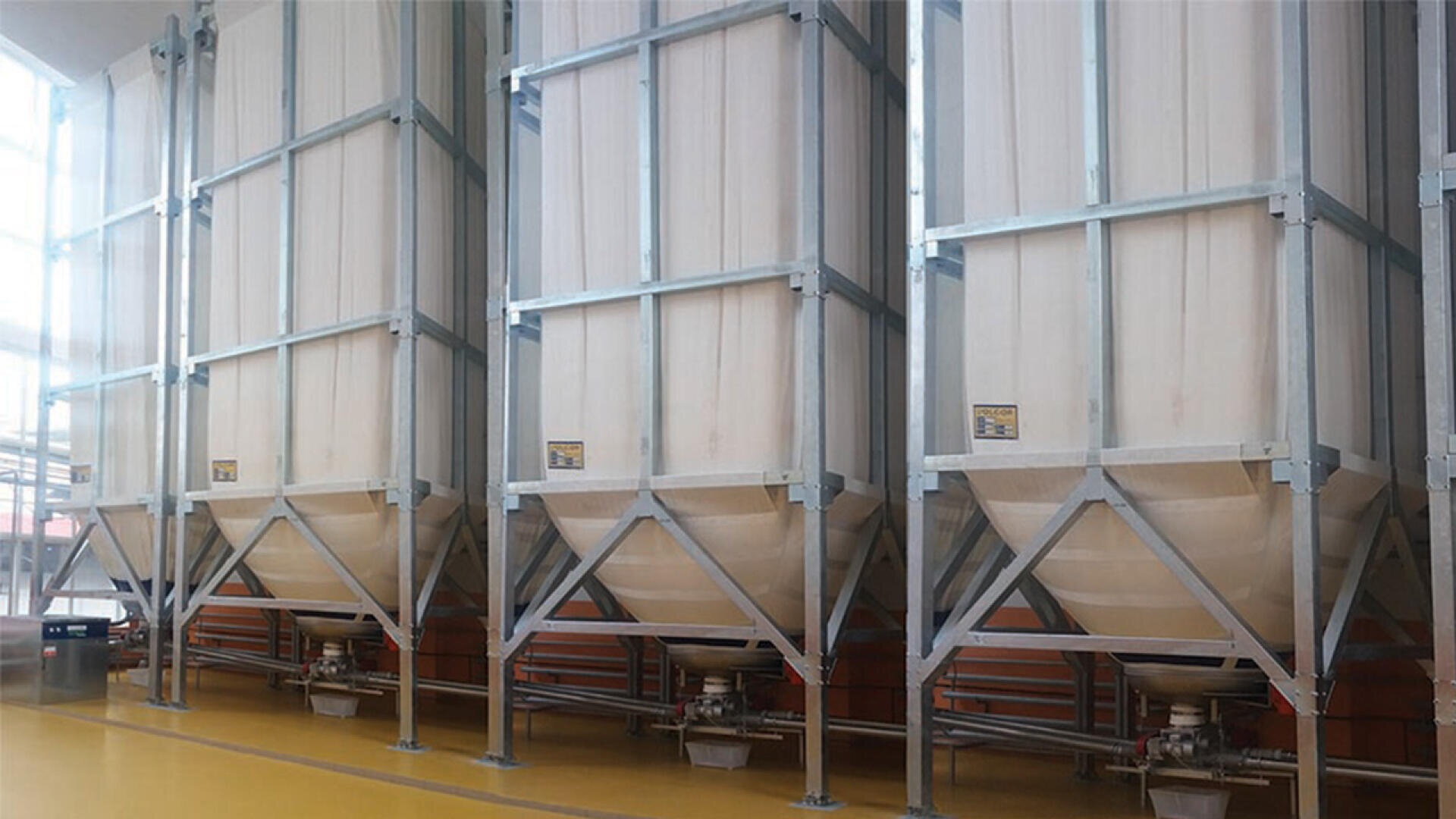 Three large industrial fabric silos with metal frames are lined up indoors on a yellow floor, used for storing bulk materials such as grains or powders. Pipes and outlets are visible at the bottom of each silo.