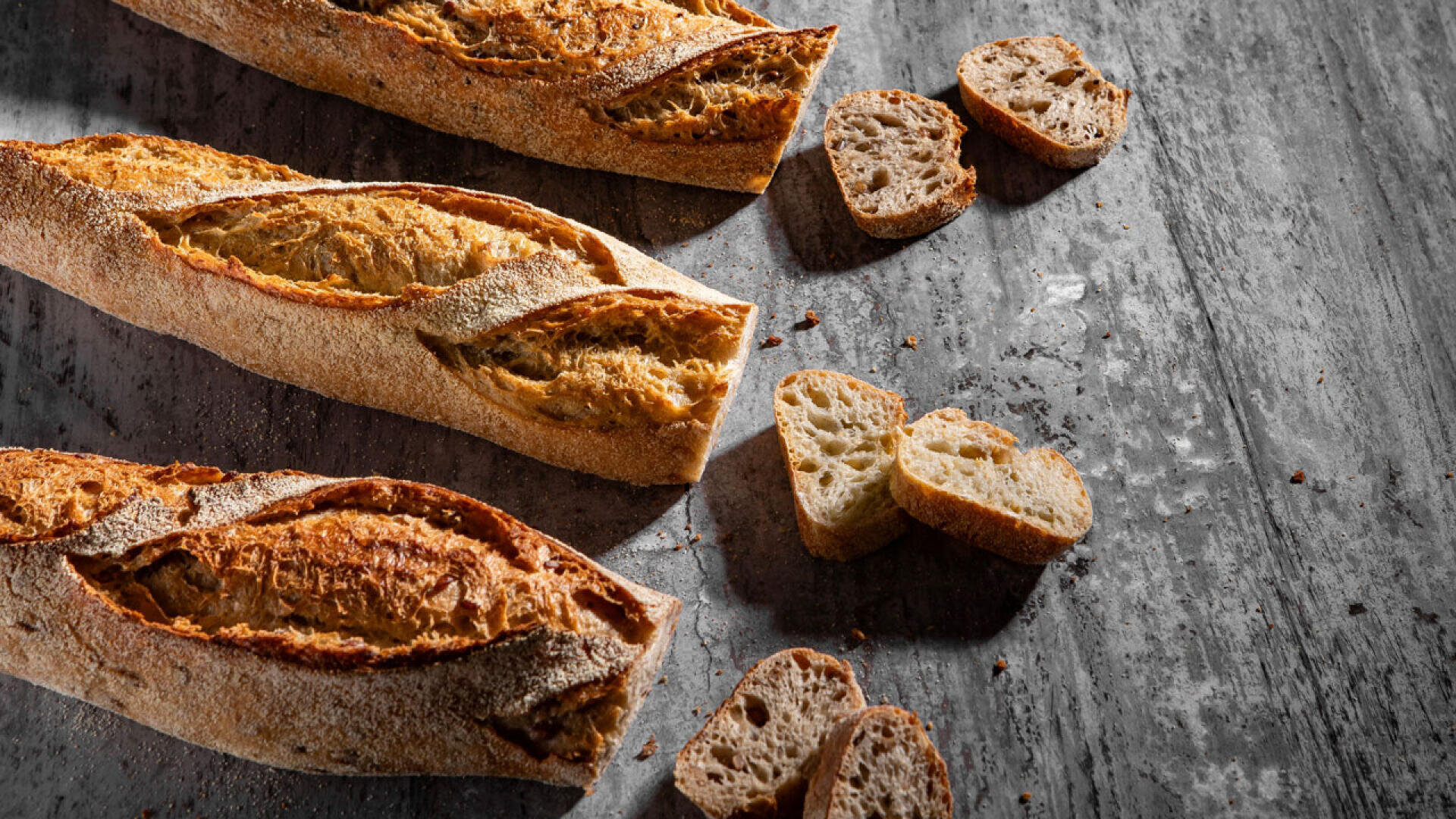 Drei knusprige Baguettes und mehrere Brotscheiben sind auf einer rustikalen grauen Fläche angeordnet, auf der Krümel verstreut sind.