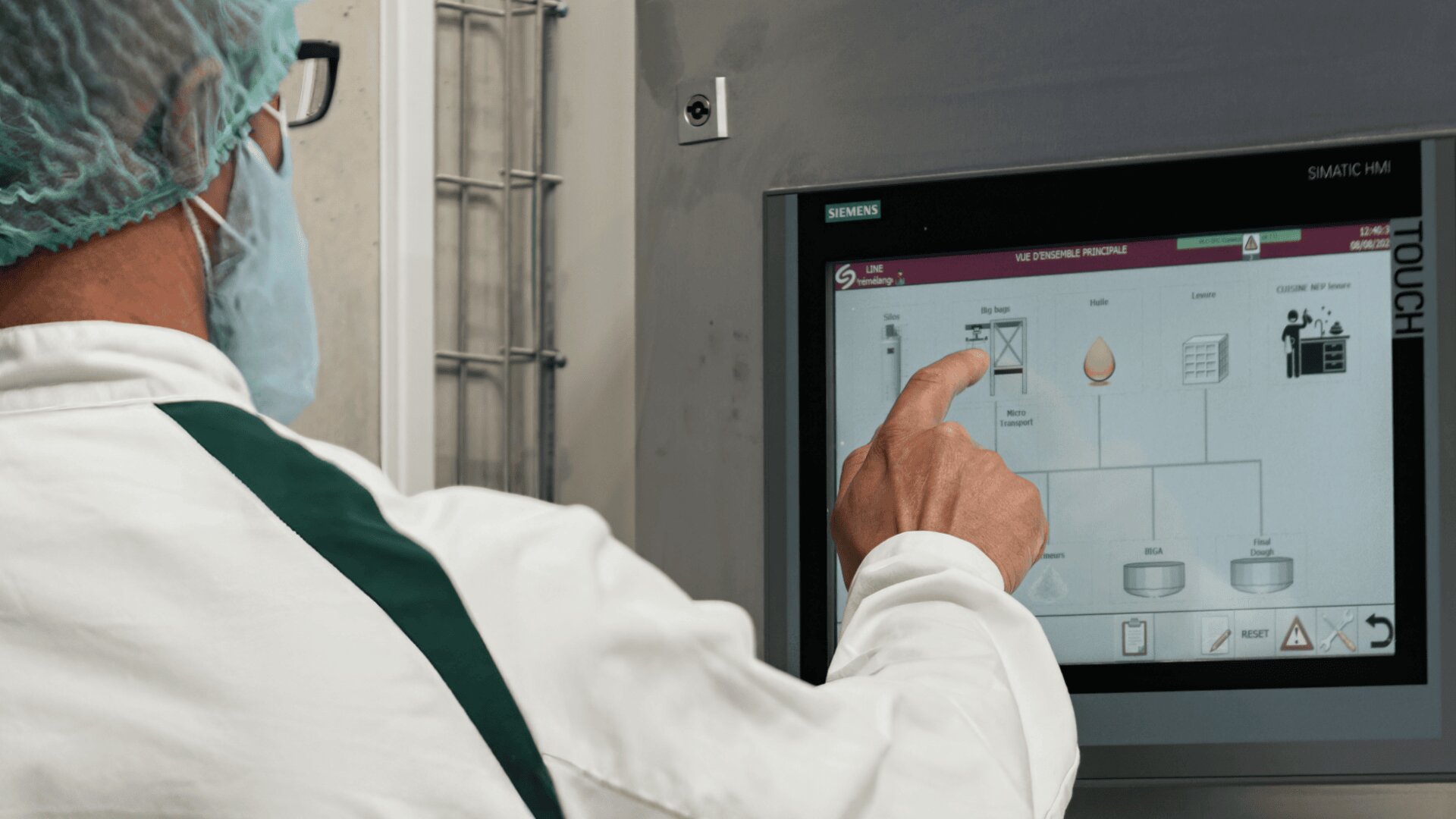 A person wearing a hairnet, face mask, and lab coat operates a Siemens touchscreen control panel displaying a process flow diagram in a laboratory or industrial setting.