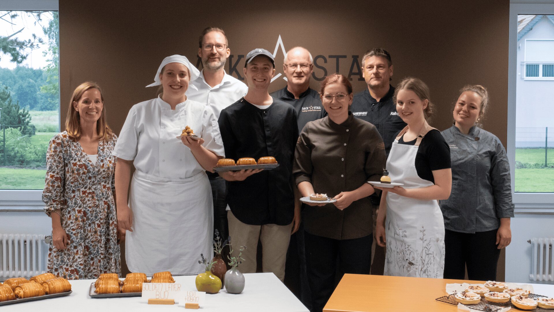 Eine Gruppe von neun Personen, einige davon in Kochuniformen, steht lächelnd in einer Küche mit Gebäck und Kuchen auf Tischen vor ihnen. Im Hintergrund sind Fenster mit Blick auf Grünflächen und eine braune Wand mit einem teilweisen Logo zu sehen.