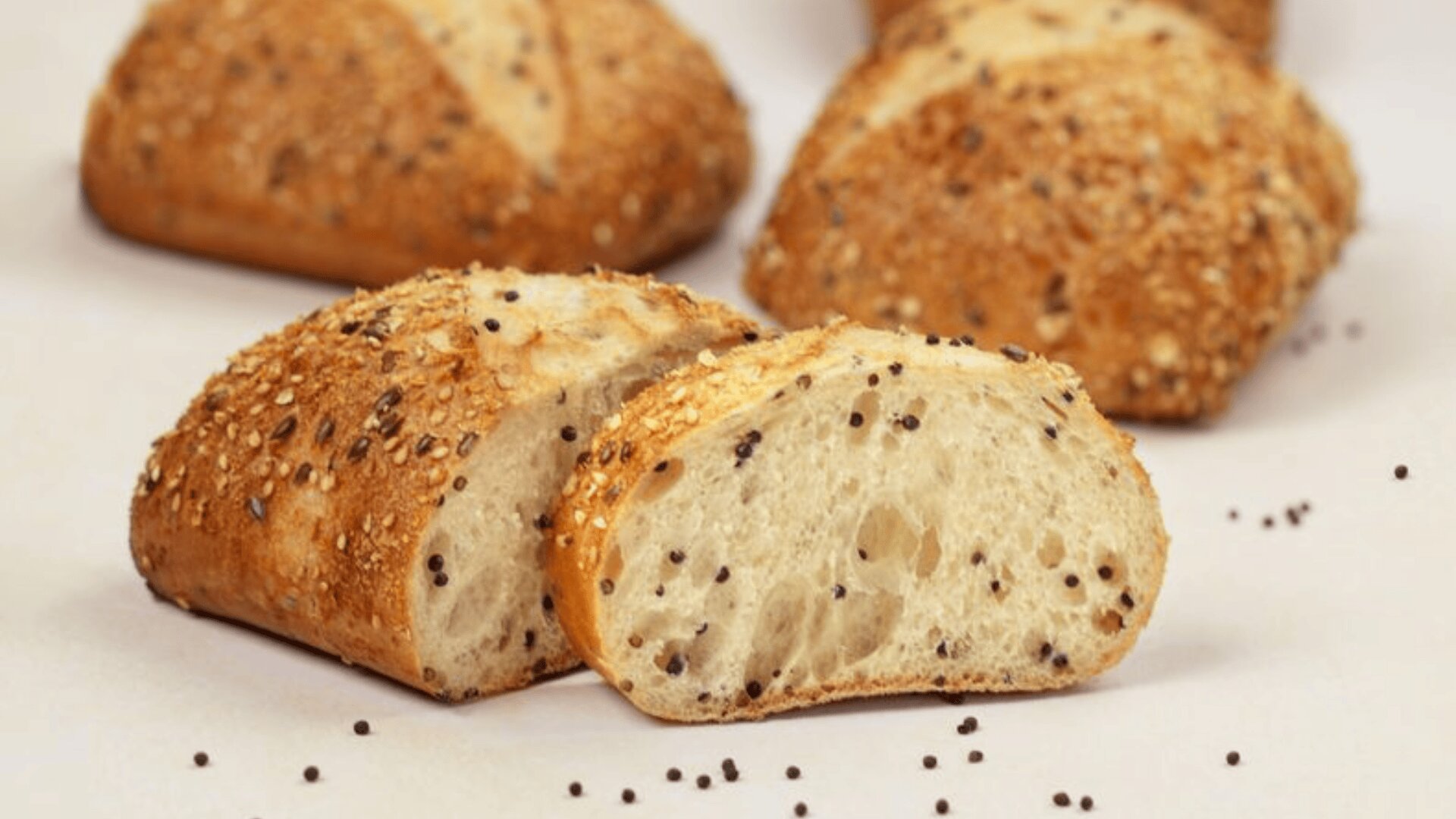 Es werden mehrere Körnerbrötchen gezeigt, wobei ein Brötchen in der Mitte aufgeschnitten ist und ein weiches, luftiges, mit Körnern übersätes Inneres zeigt. Die Kruste ist goldbraun und mit gemischten Samen bedeckt, und einige Samen sind auf der weißen Oberfläche verstreut.