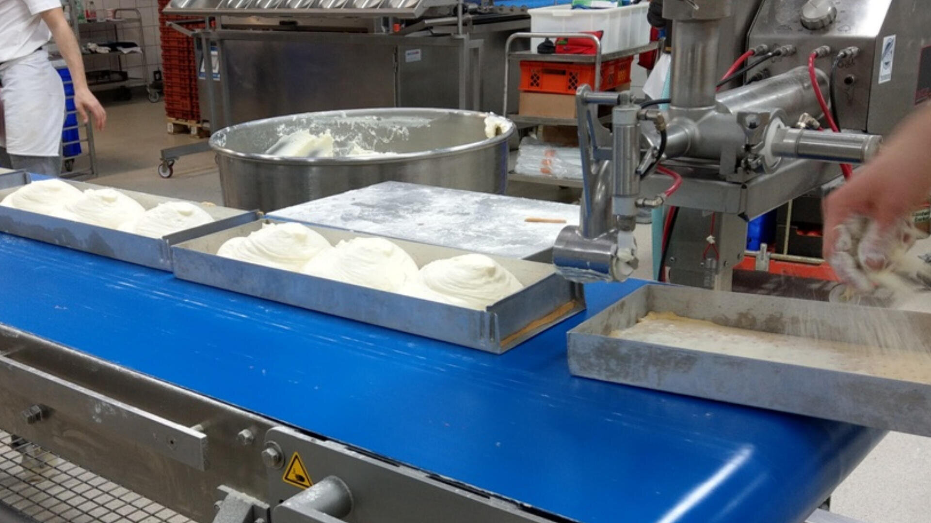 Eine Bäckereifertigungsstraße mit Metallschalen mit Teig auf einem blauen Förderband, eine industrielle Maschine, die den Teig ausgibt, und Arbeiter, die die Schalen in einer sauberen, hellen Anlage bearbeiten.