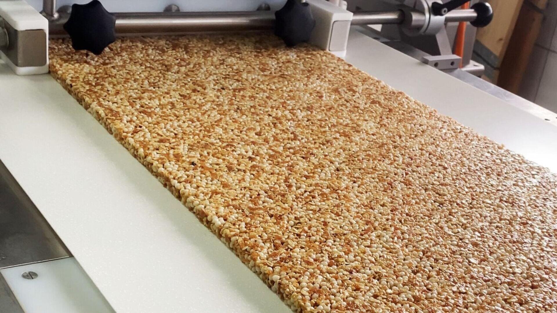 Eine Maschine presst und formt eine große, flache Platte aus gepufftem Reis oder Getreide, die mit Sirup verbunden ist, wahrscheinlich für die Herstellung von Snack-Riegeln. Die Mischung ist goldbraun und gleichmäßig strukturiert.