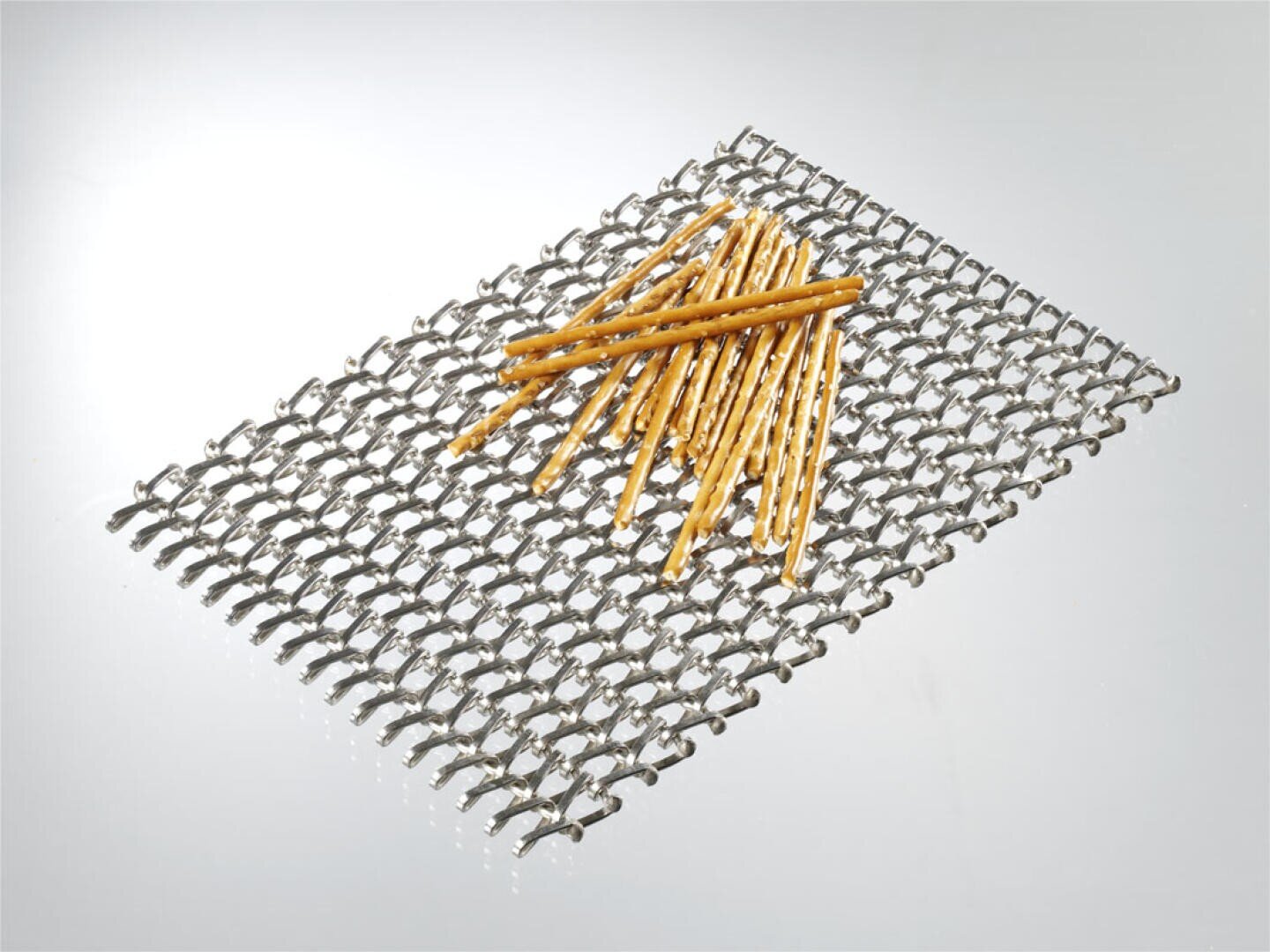 Ein Stapel Laugenstangen liegt auf einem rechteckigen Metallgitter eines Förderbandes vor einem schlichten, hellen Hintergrund.