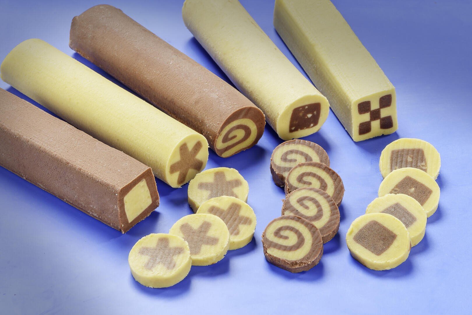 Verschiedene Stämme gemusterten Keksteigs, einige mit Schokoladen- und einige mit Vanillegeschmack, werden in Scheiben geschnitten, um Muster wie Spiralen, Xs und Schachbrettmuster auf einer blauen Oberfläche zu zeigen. Die Scheiben zeigen die verschlungenen Formen im Inneren.