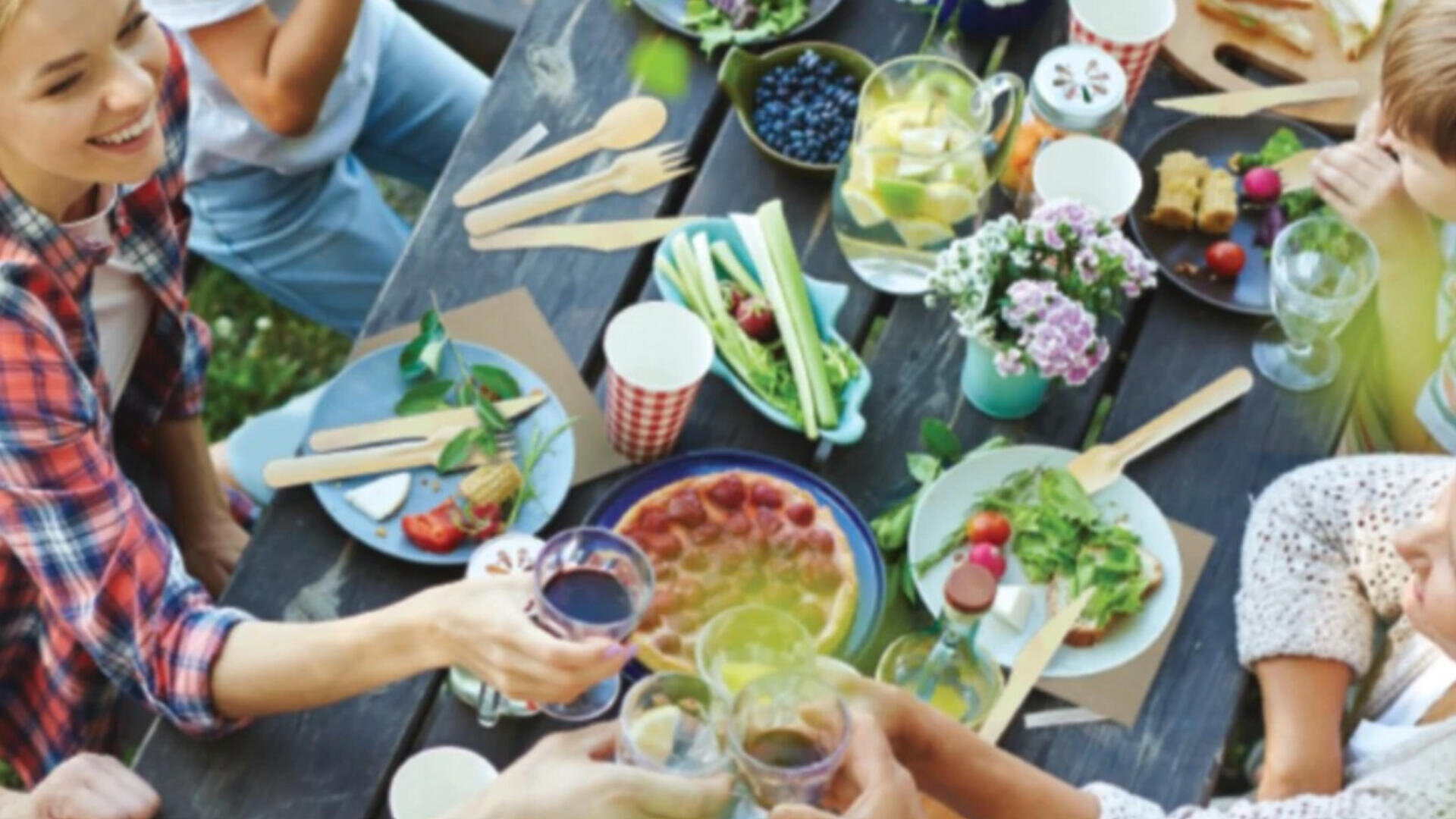 A group of people gather around a picnic table filled with food, drinks, flowers, and wooden utensils, toasting with glasses in a cheerful outdoor setting.