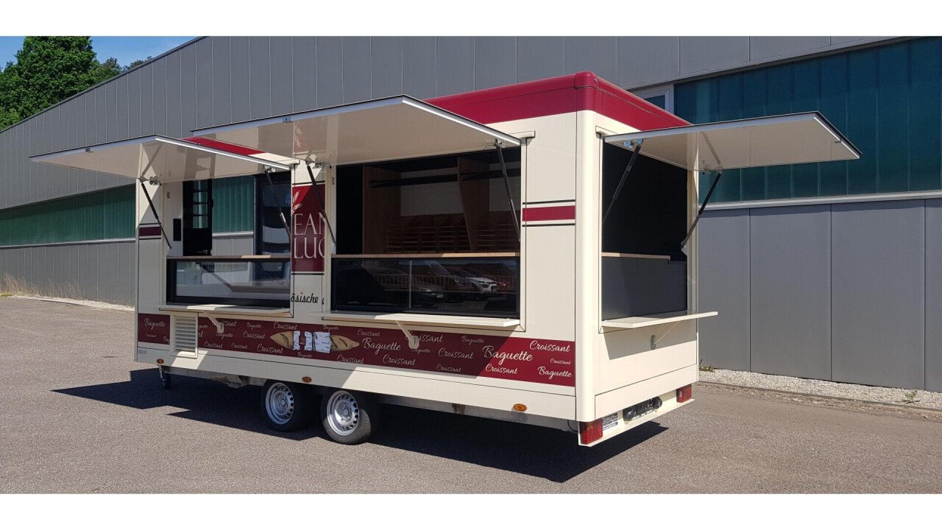 A cream and red food trailer with open serving windows on three sides, standing on pavement near a large industrial building with green-tinted glass panels. The trailer has display graphics showing baguettes and pastries.
