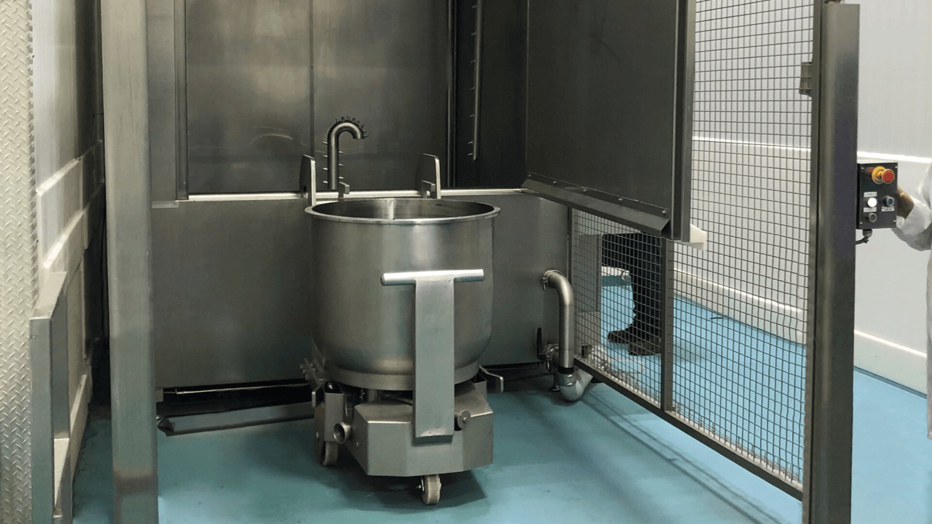 Ein großer industrieller Mischkessel aus rostfreiem Stahl auf Rädern befindet sich in einem Metallkäfig mit einem offenen Tor. Eine Person steht in der Nähe und drückt einen roten Knopf auf einem Bedienfeld. Der Raum hat einen blauen Fußboden und weiße Wände.