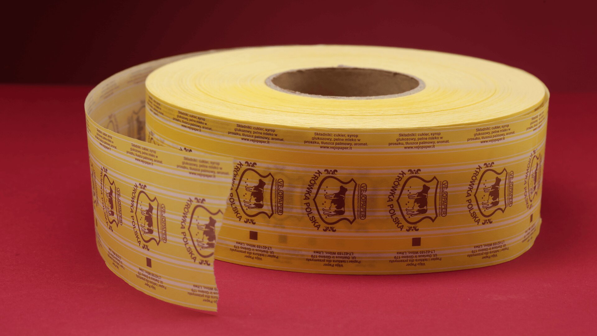 Eine große Rolle transparenter gelber Verpackungsfolie mit aufgedruckten braunen und gelben Logos und Texten, die leicht aufgerollt ist, befindet sich auf einer roten Fläche vor einem roten Hintergrund.