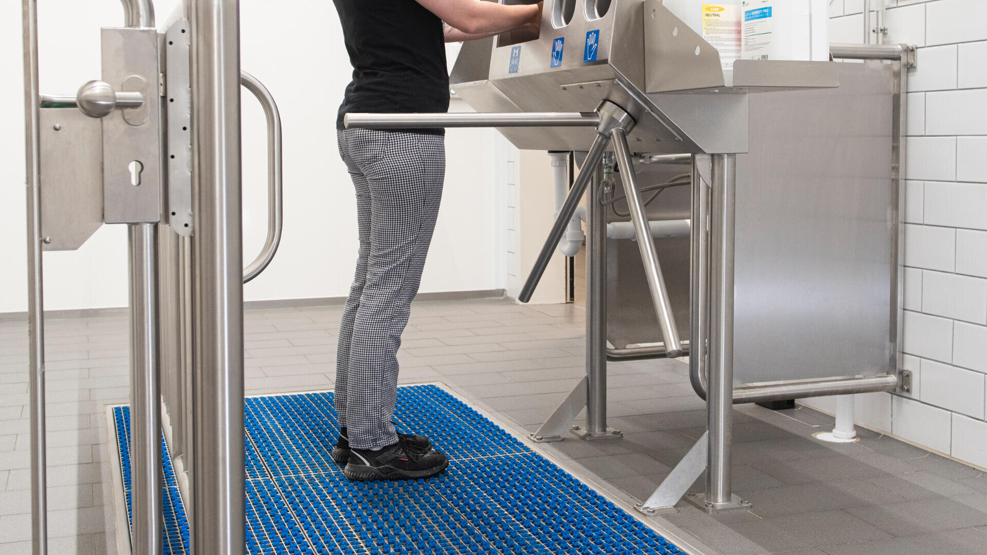 Person standing on a blue mat, using a stainless steel hygiene station with turnstile in a clean, tiled facility. Only the lower half of the person is visible.
