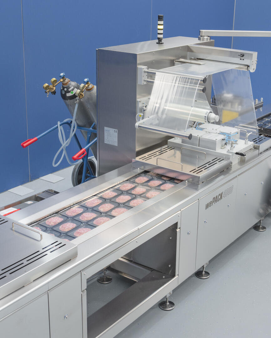 Eine Lebensmittelverpackungsmaschine wickelt mehrere Schalen mit rohen Hamburger-Patties in Plastikfolie in einer sauberen, industriellen Umgebung ein. Im Hintergrund sind Gasflaschen und blaue Wände zu sehen.