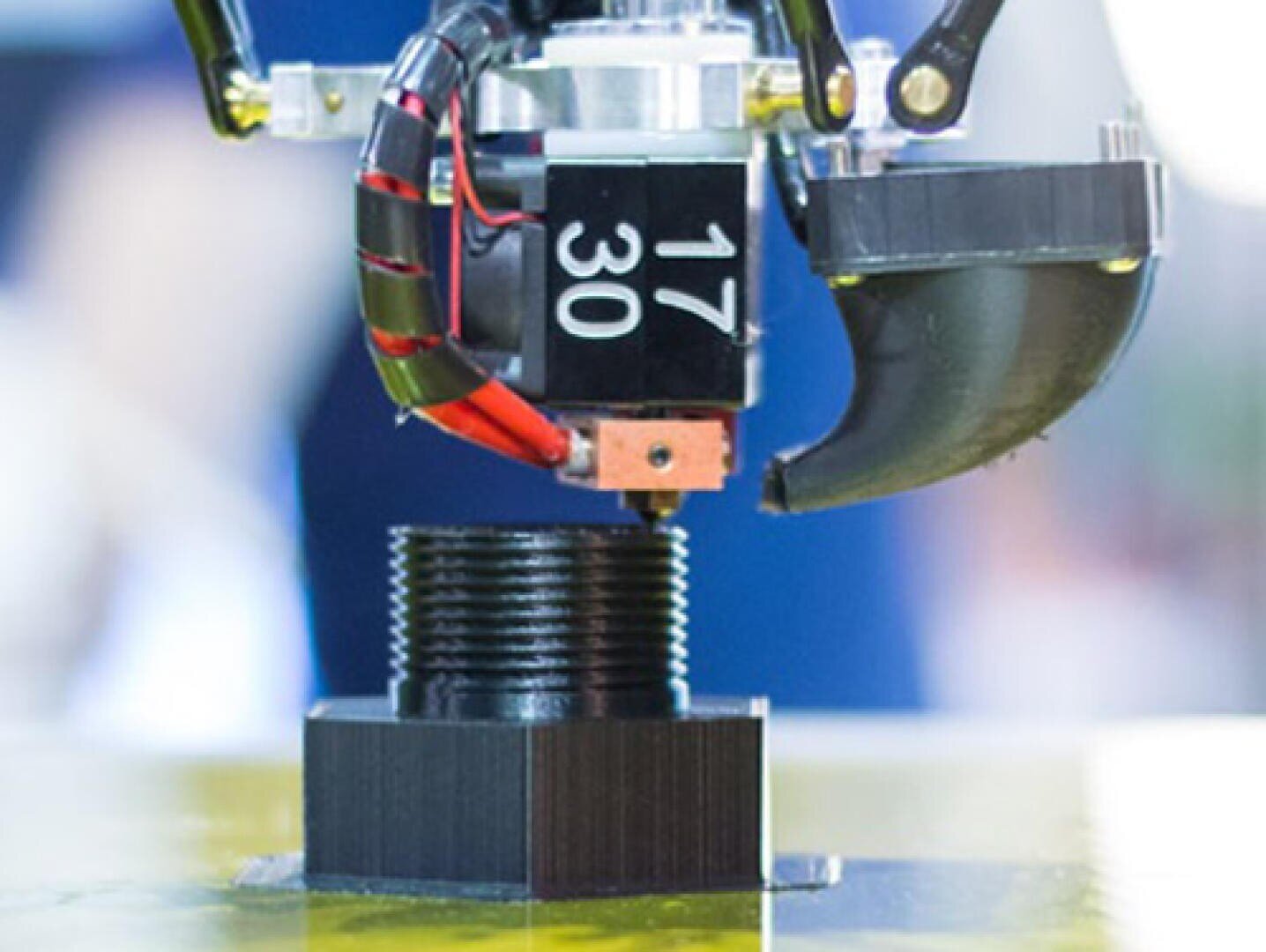 A close-up of a 3D printer nozzle printing a black, cylindrical, threaded object layer by layer on a yellow platform. Red and black wires are visible near the nozzle.