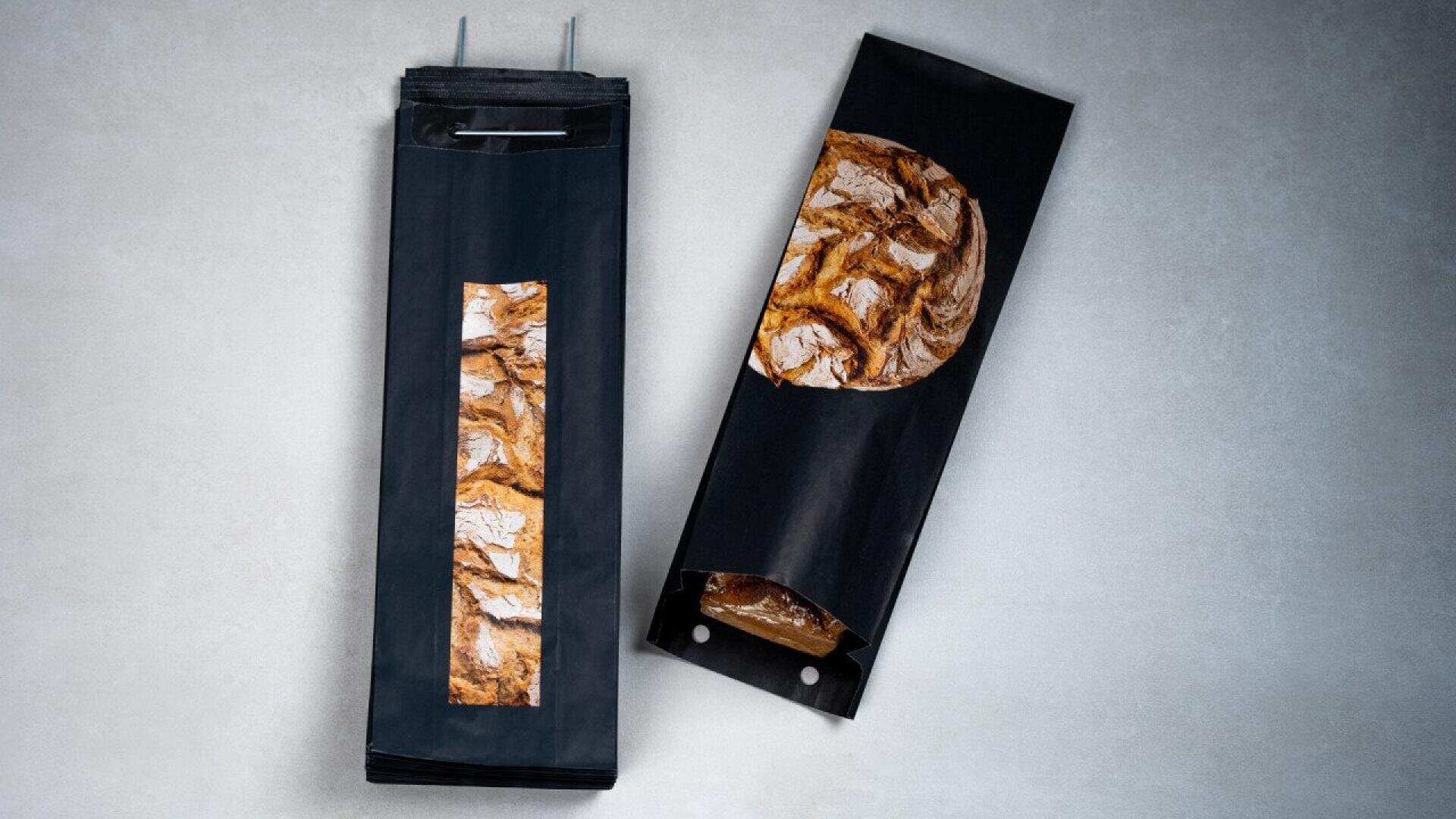 Two black paper bread bags with a window showing the crusty loaf inside; one bag stands upright while the other lies flat, partially revealing the round bread.