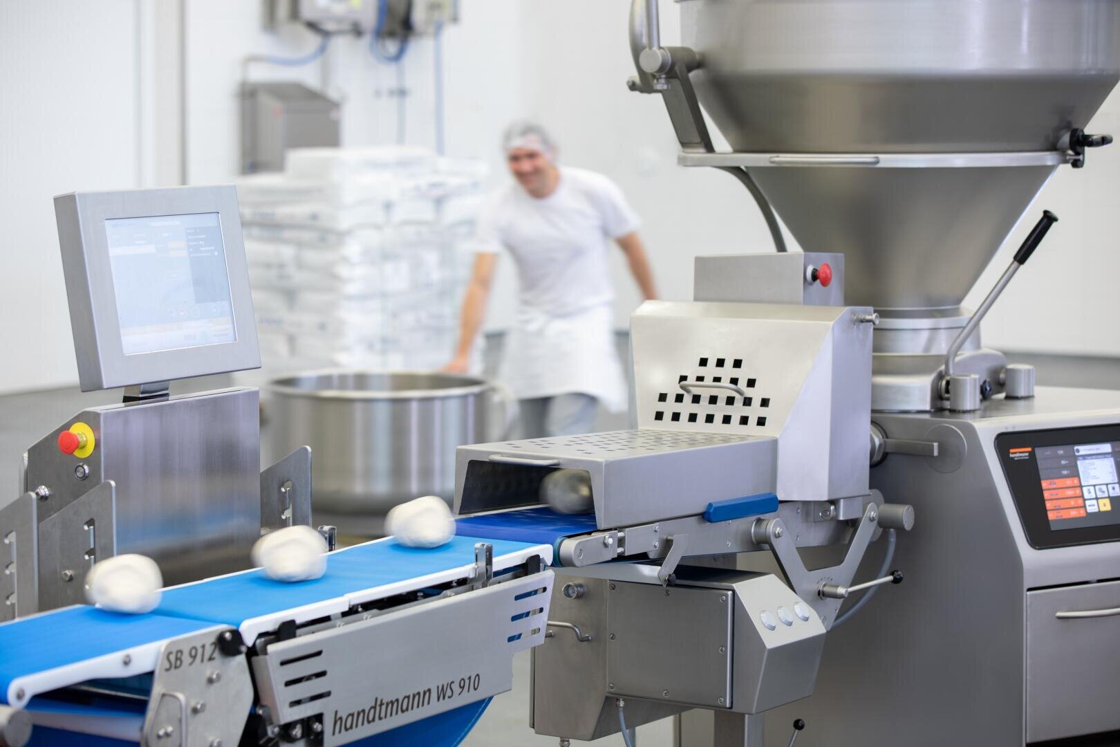 Eine Lebensmittelverarbeitungsmaschine legt Teigkugeln auf ein blaues Förderband in einer sauberen Industrieküche. Im Hintergrund ist eine Person in weißer Arbeitskleidung und mit Haarnetz unscharf zu erkennen.
