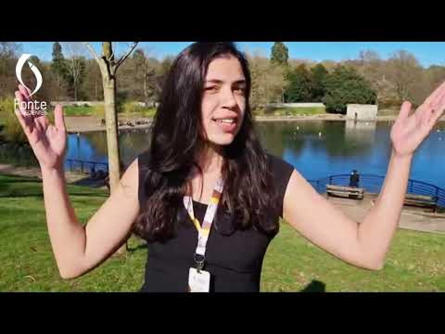 A woman with long dark hair stands outside near a lake, raising her arms with a smile. She is wearing a black top and a lanyard. Trees, grass, benches, and water are visible in the background on a sunny day.
