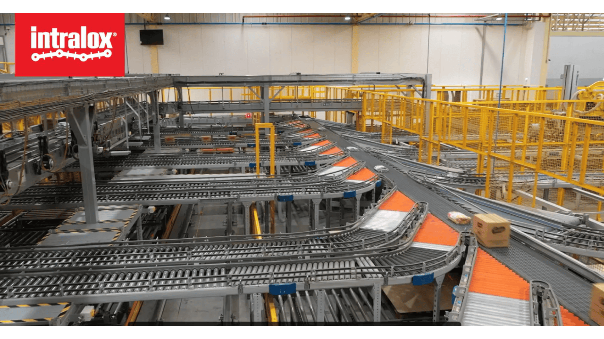 Ein Lager mit automatisierten Förderbändern zum Transport von Paketen, mit gelben Sicherheitsgeländern und dem Intralox-Logo in der oberen linken Ecke.