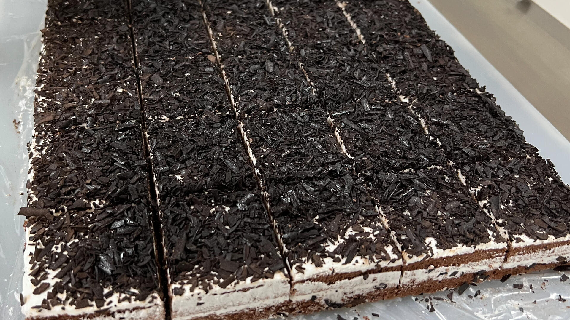 A tray of rectangular chocolate cakes with white cream and dark chocolate shavings on top, cut into even squares and arranged in rows.