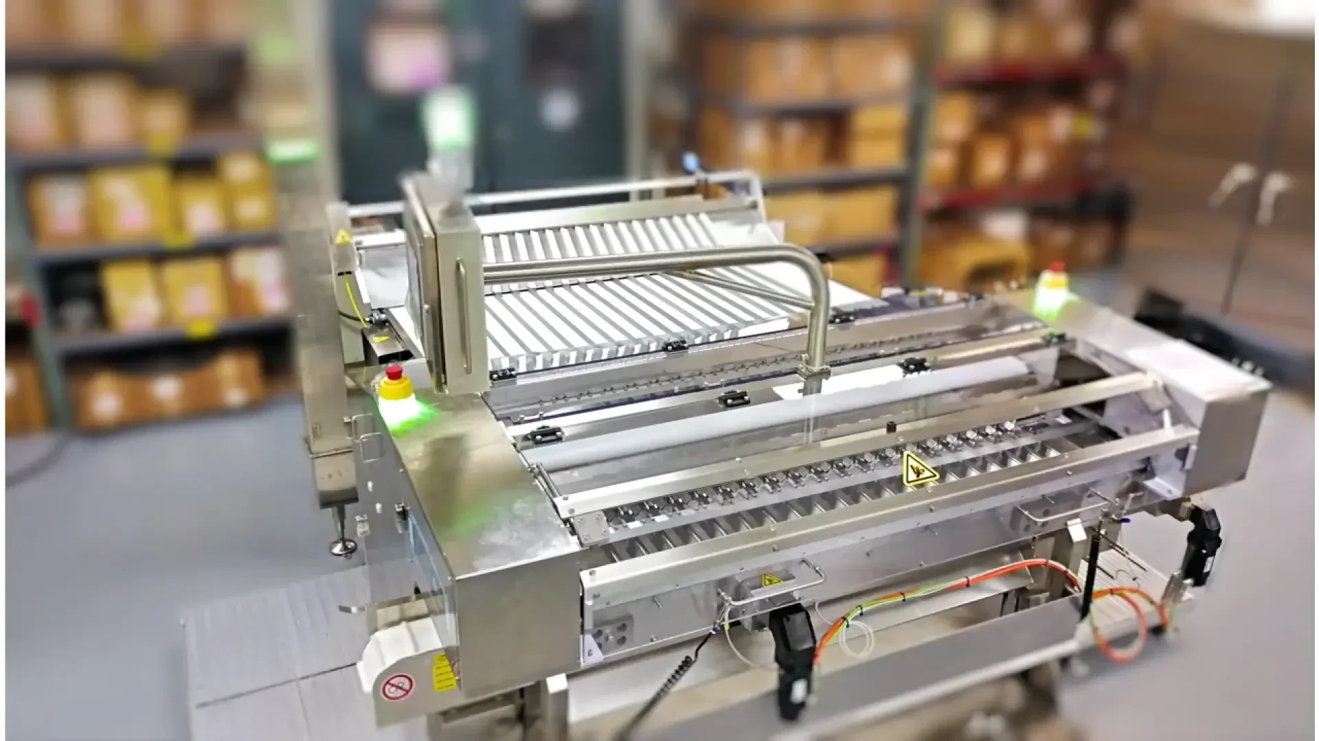 Industrial packaging machine with metallic rollers, safety guards, and control buttons, situated in a warehouse with shelves of brown boxes in the blurred background.