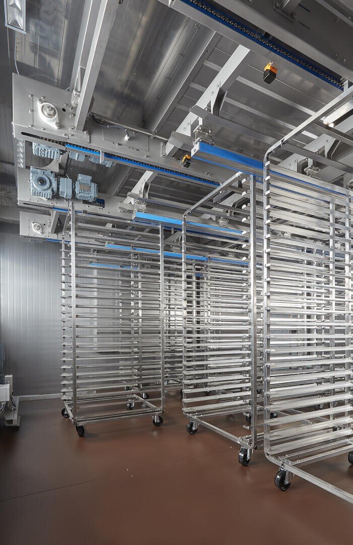 Rows of empty metal bakery racks with wheels are lined up in a large industrial kitchen or food processing facility, featuring stainless steel surfaces, a brown floor, and exposed ceiling machinery.