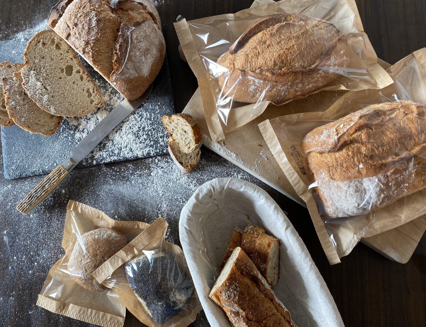 A table with several loaves of crusty bread, some sliced and some in paper bags, a bread knife, a dusting of flour, and a bread basket holding a few slices.