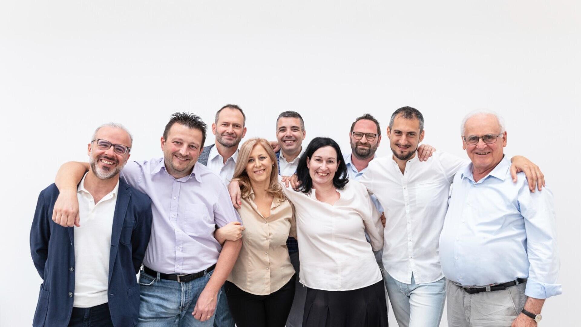 A group of nine adults, men and women, stand together smiling against a plain white background. They are casually dressed and appear friendly, with some linking arms or placing hands on each others shoulders.