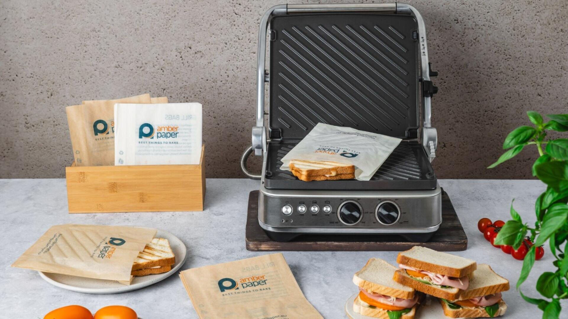 Eine Sandwichpresse mit einem Sandwich darin steht auf einer Arbeitsplatte. Daneben liegen eingepackte und ausgepackte Sandwiches, braune Sandwichtüten, frisches Gemüse und ein Holzständer mit Papiertüten mit der Aufschrift "Amber Paper".
