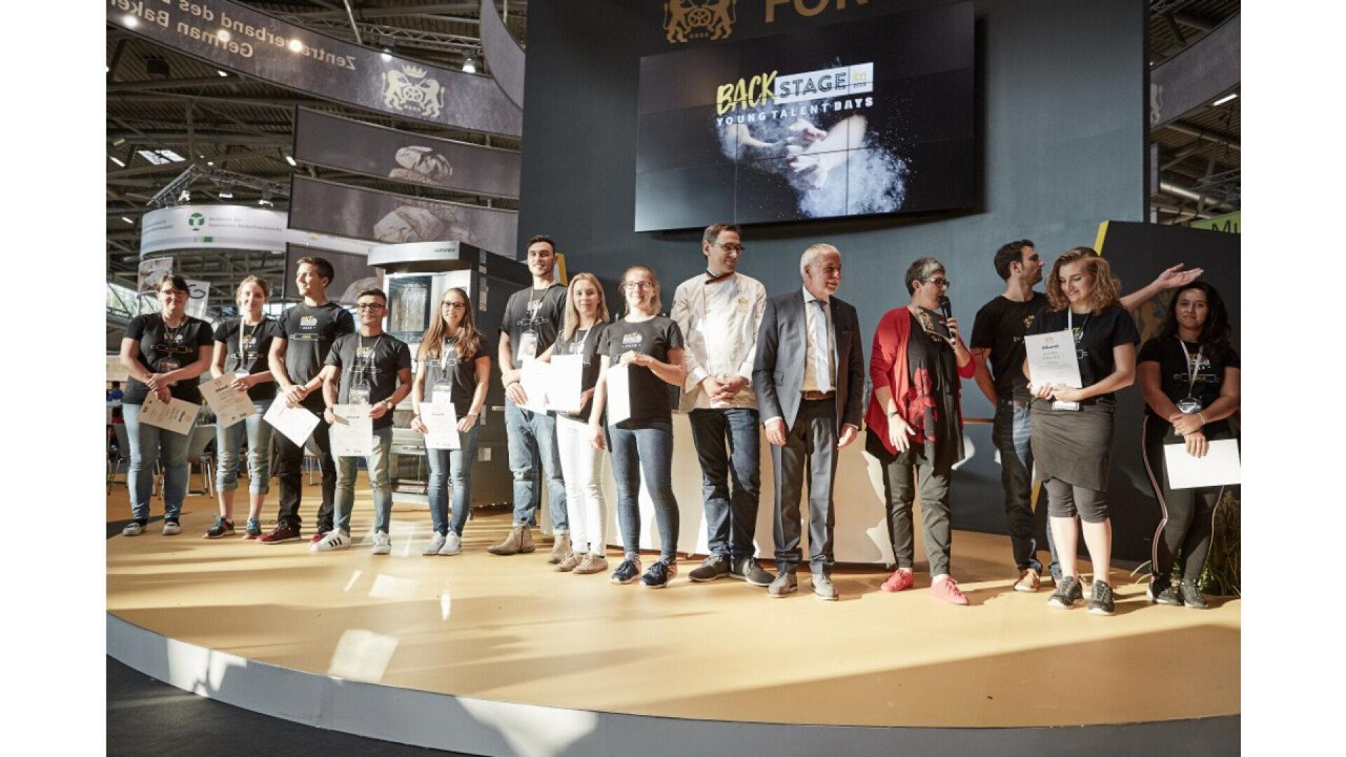 A group of people stand on a stage, some holding certificates. Behind them is a large screen displaying Backstage with other text and graphics. The setting appears to be an indoor event or exhibition.