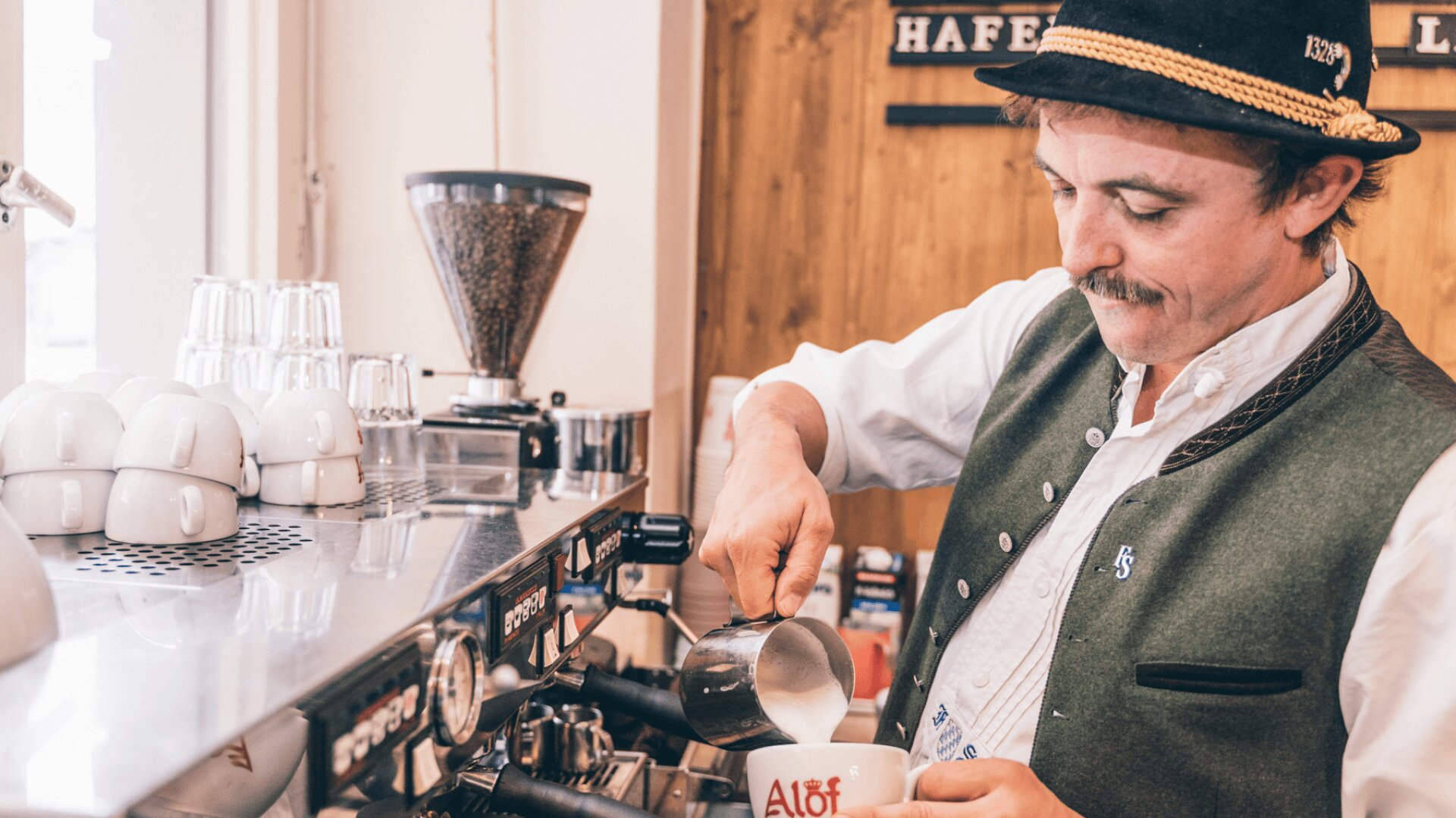 Ein Barista in bayerischer Tracht gießt an einer Espressomaschine in einem gemütlichen Café dampfende Milch in eine Kaffeetasse. Auf dem Tresen sind Stapel von weißen Tassen und eine Kaffeemühle zu sehen.
