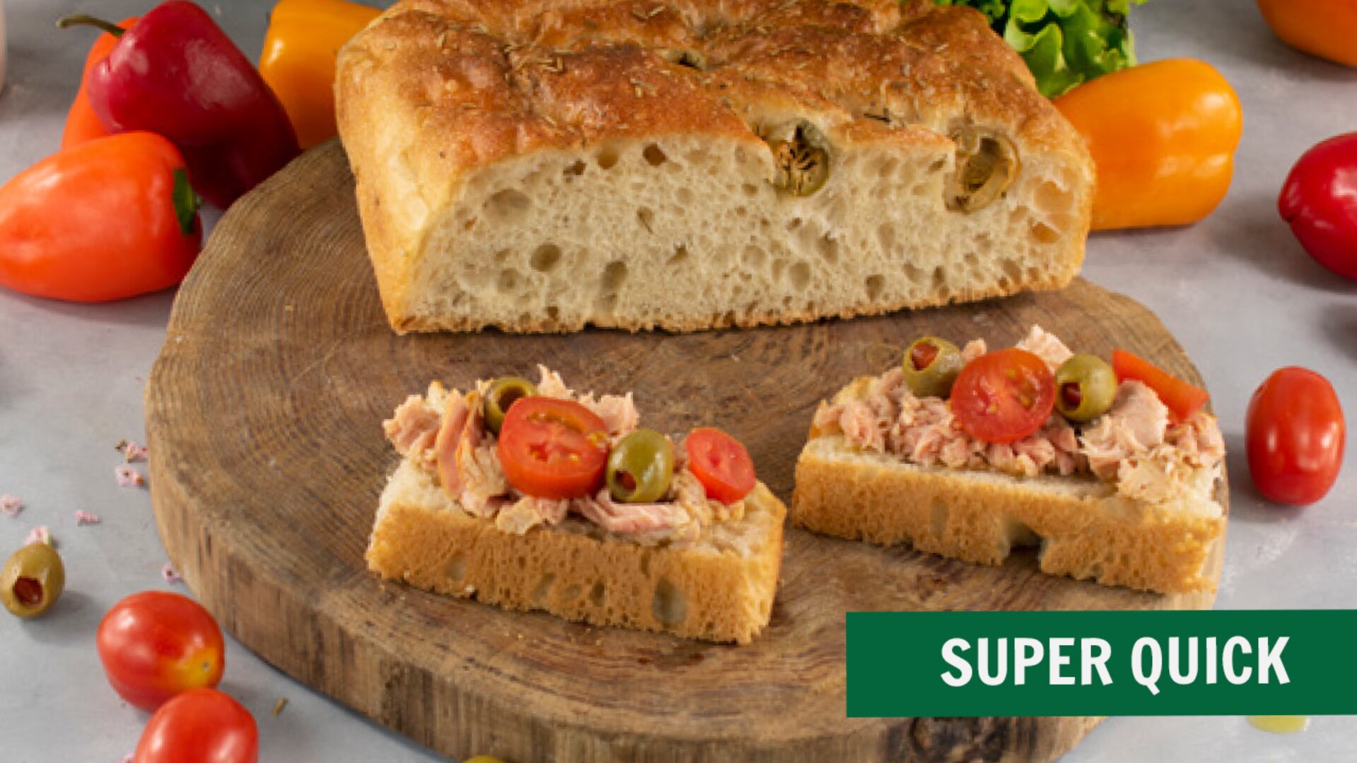 A wooden board with focaccia bread, some topped with tuna, sliced olives, and cherry tomatoes. Colorful bell peppers and tomatoes are scattered around. A green label reads SUPER QUICK.
