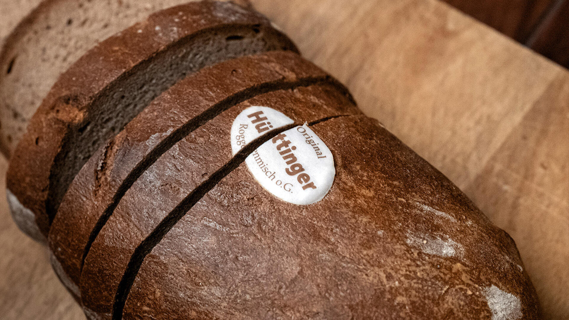 Ein Laib geschnittenes Schwarzbrot auf einer Holzoberfläche, obenauf ein Etikett mit der Aufschrift „Original Hüttinger Roggenschmisch OG“.