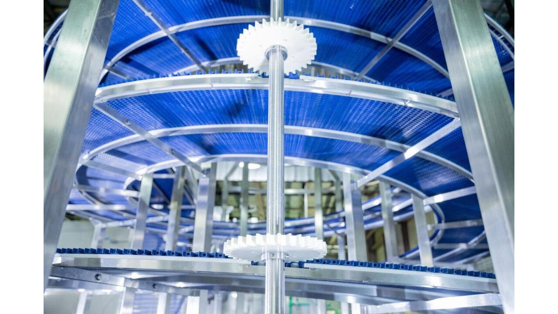 Nahaufnahme eines Spiralfördersystems mit blauen Maschengurten, Metallschienen und weißen Kunststoffzahnrädern, das in der Industrie oder Lebensmittelverarbeitung eingesetzt wird. Die Perspektive zeigt die kreisförmige Struktur von unten.