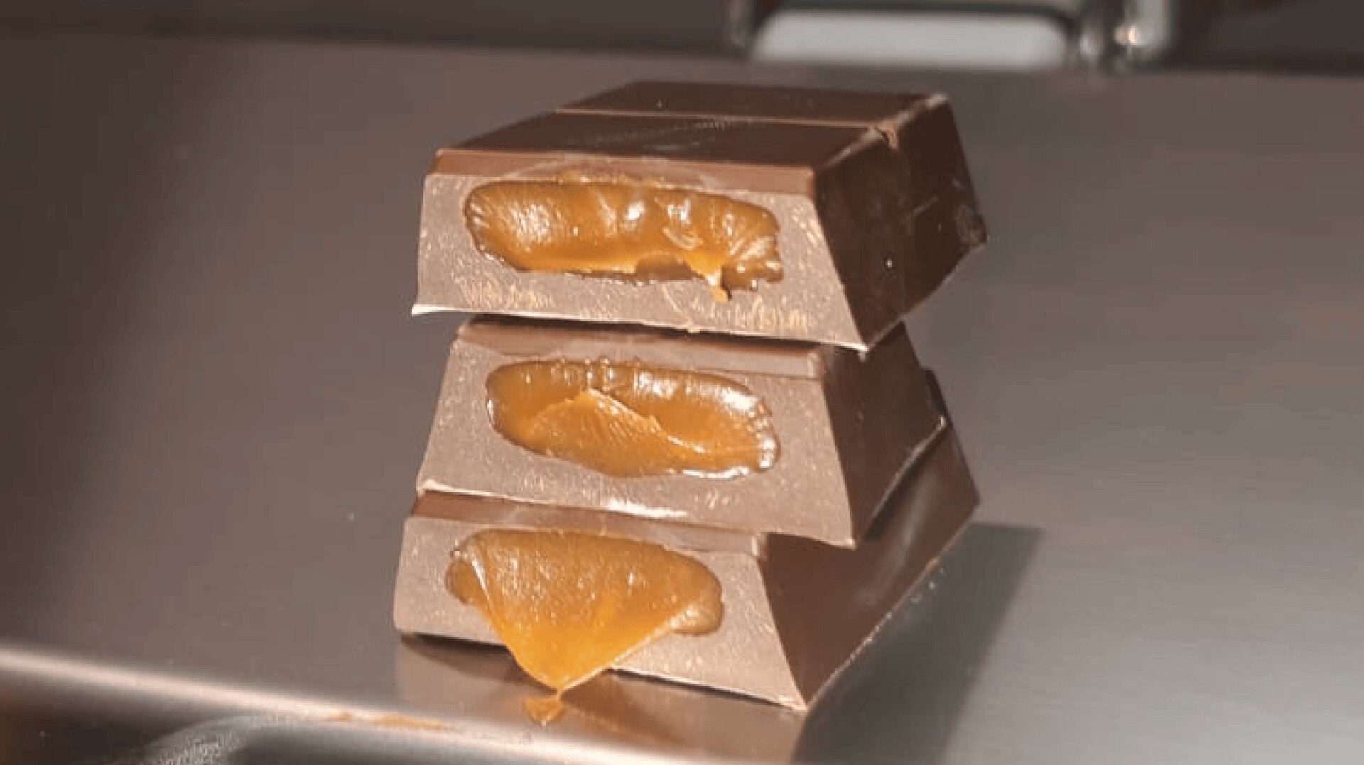 Three pieces of chocolate stacked on top of each other, each with a gooey caramel filling visible and slightly oozing out. The chocolates are placed on a smooth, metallic surface.