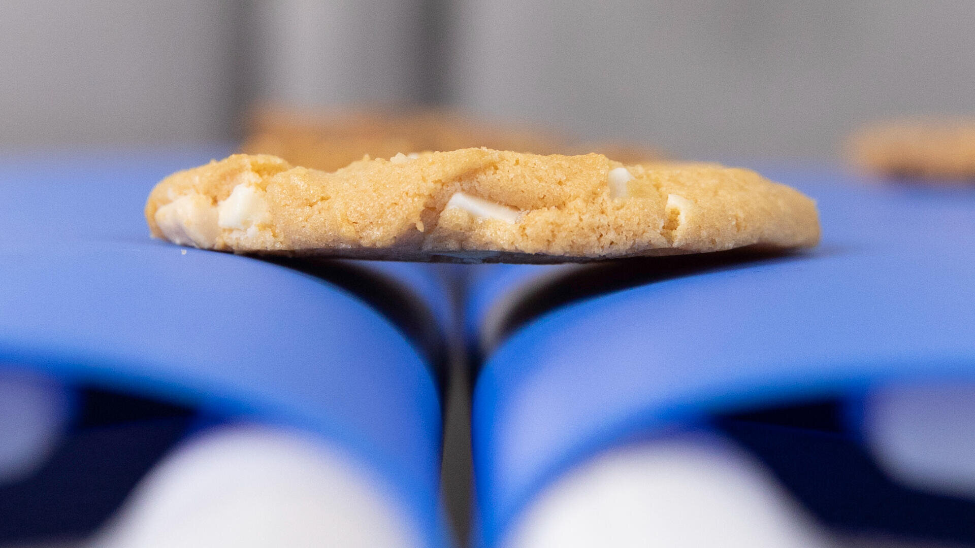 A close-up of a cookie with white chocolate chips resting on two parallel blue surfaces, with a blurred background and another cookie visible further away.
