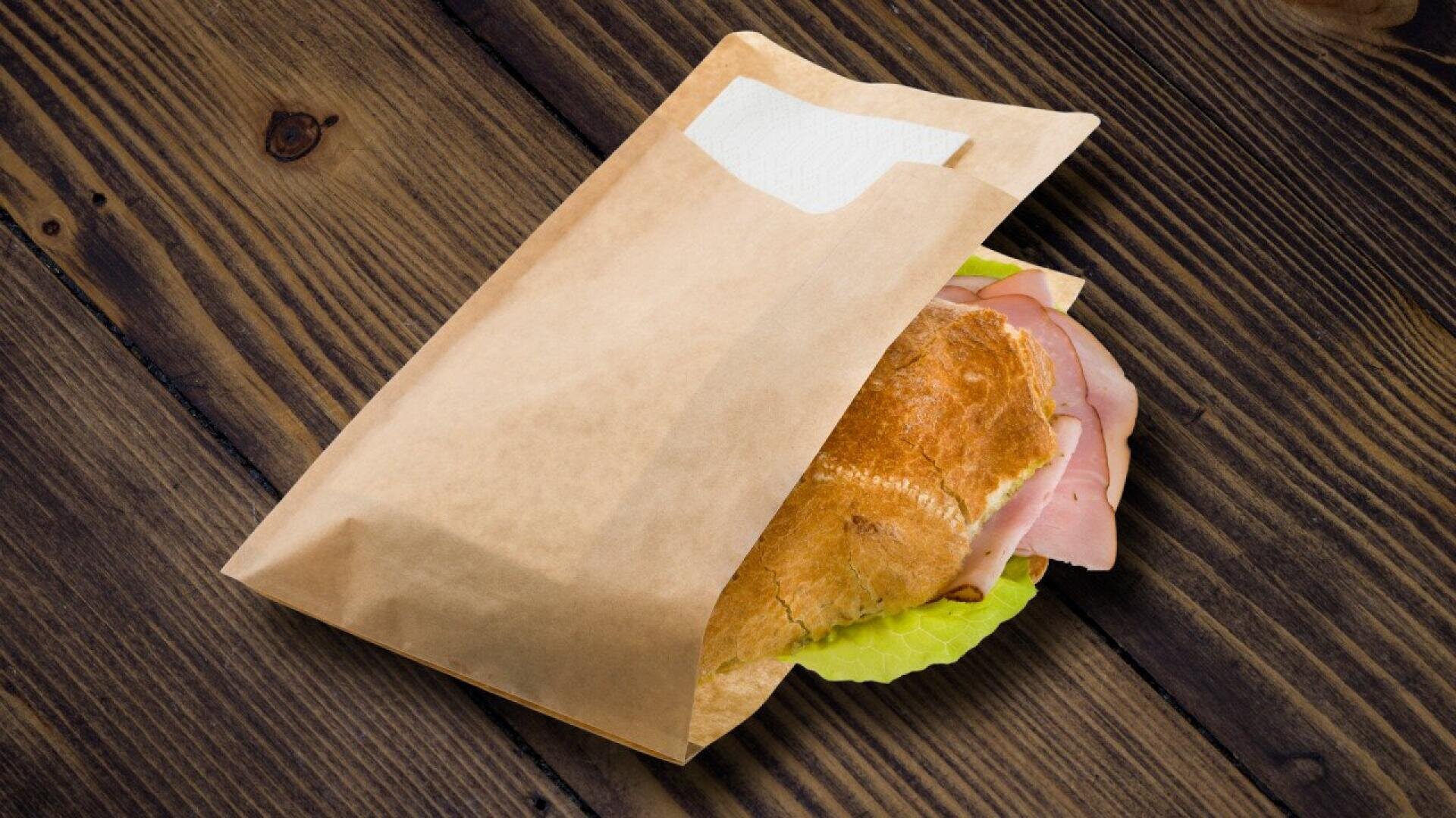 Ein Croissant-Sandwich mit Schinken und Salat, teilweise in eine braune Papiertüte mit einer Serviette eingewickelt, auf einem Holztisch.